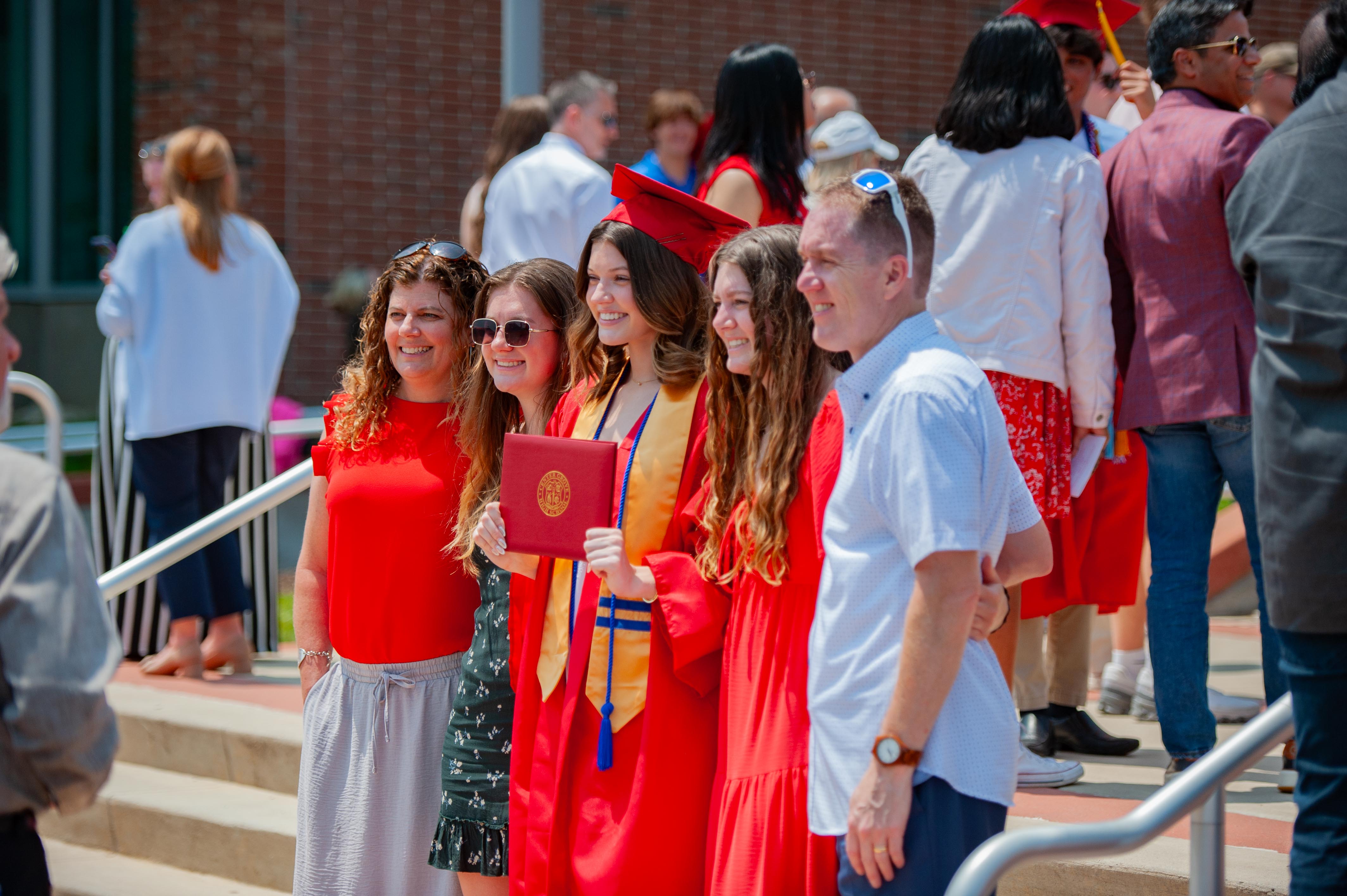 A group of smiling people in graduation attire pose for a photo on steps in front of a brick building with other attendees in the background.
