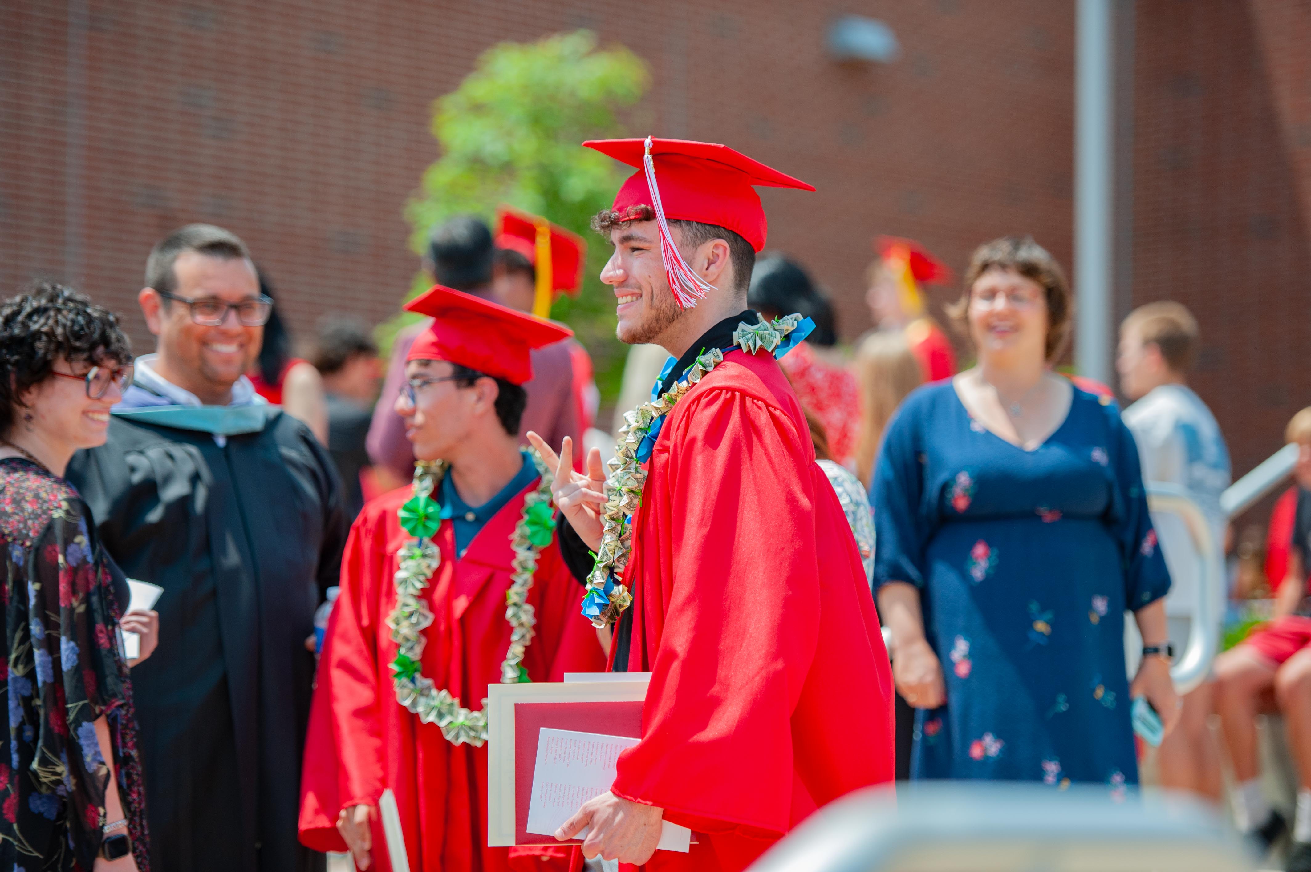 Graduates in red gowns and caps smile and pose with leis in the foreground, while a brick building and other attendees form the background.