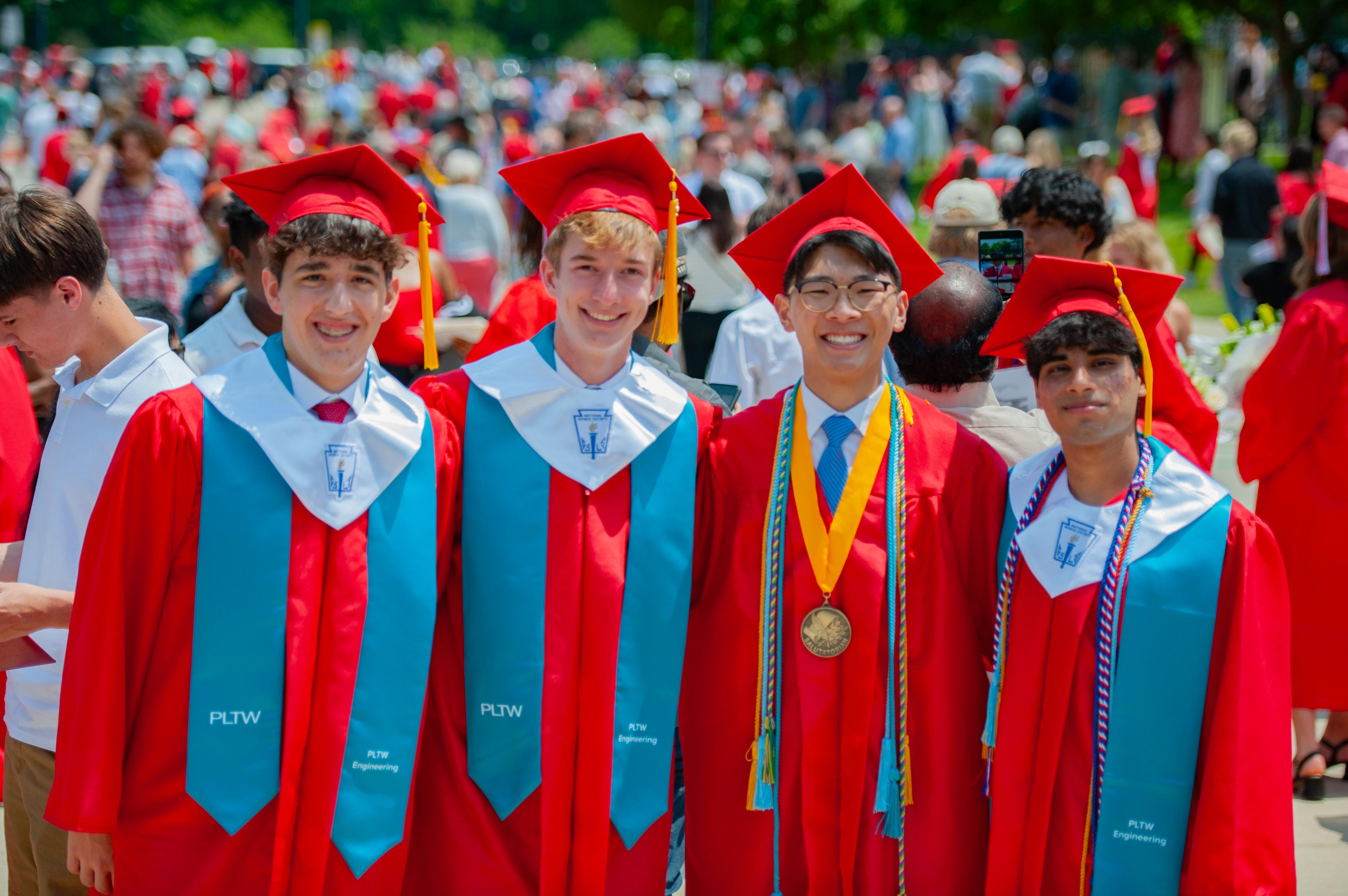 Four smiling graduates in red caps and gowns stand in the foreground, while a blurred crowd of similarly dressed individuals fills the background.
