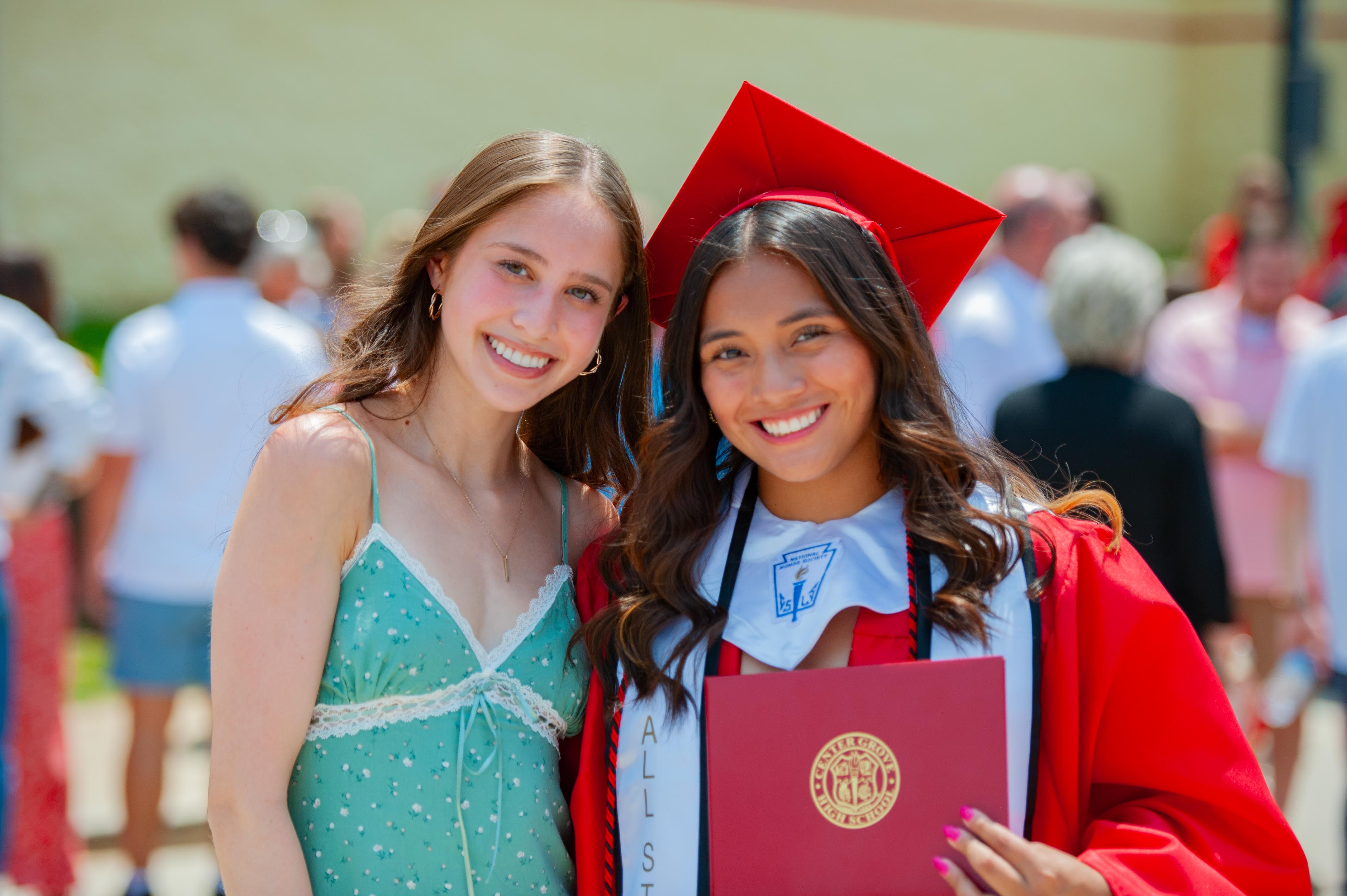 Two smiling young women, one in a graduation cap and gown holding a diploma, pose in the foreground with a blurred crowd of people in the background.