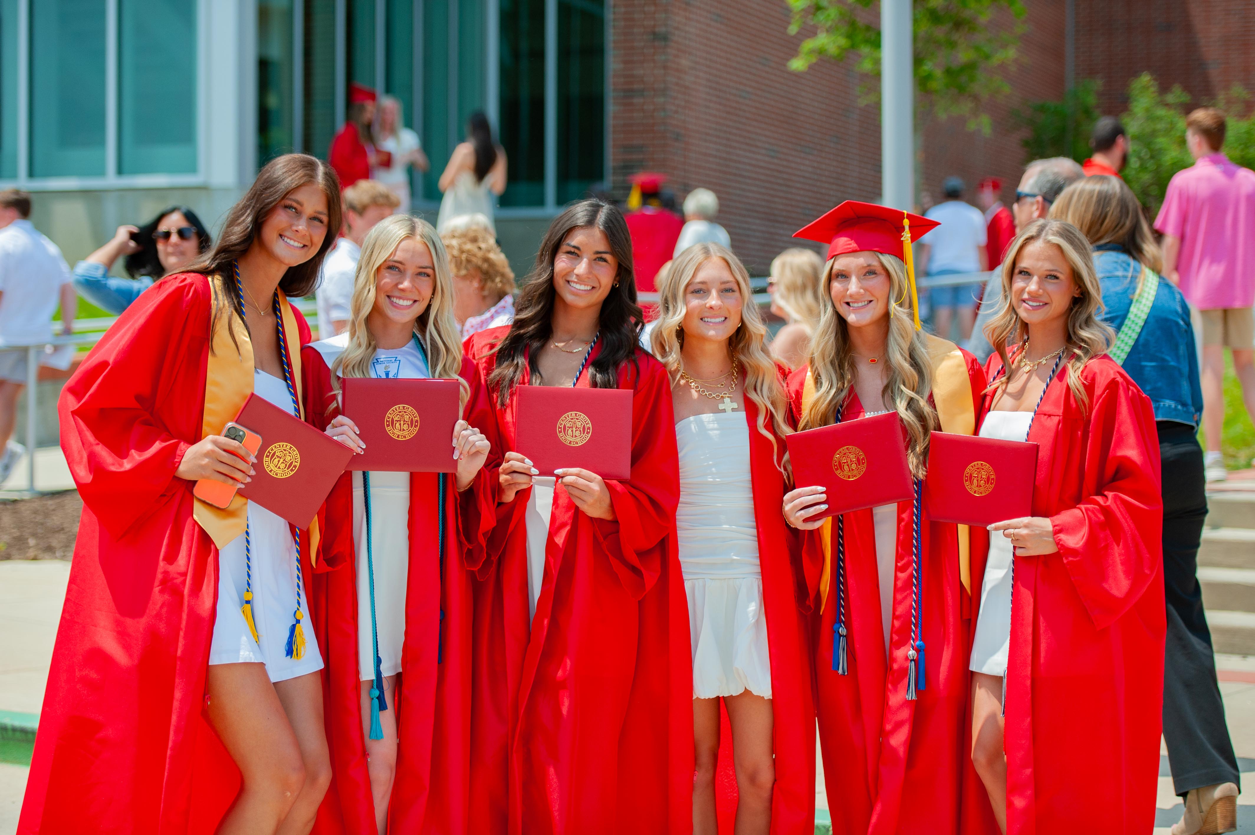 Six smiling young women in red graduation gowns and white dresses hold diplomas in front of a building with other graduates in the background.