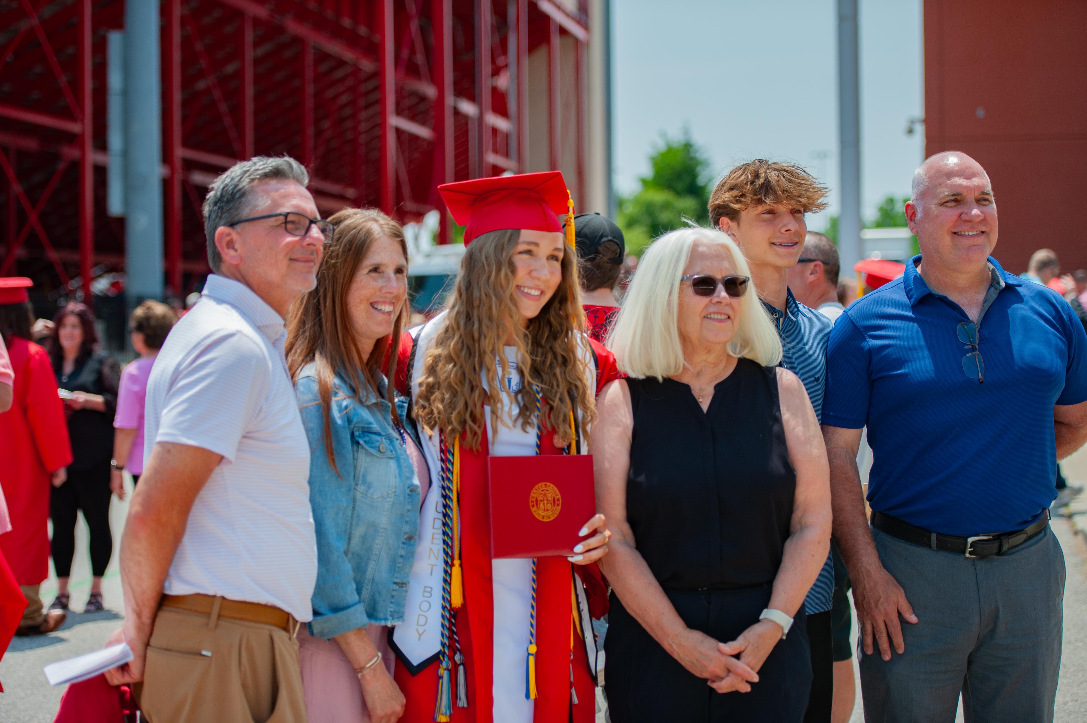 A graduating student in a red cap and gown poses with family in front of a red stadium structure.