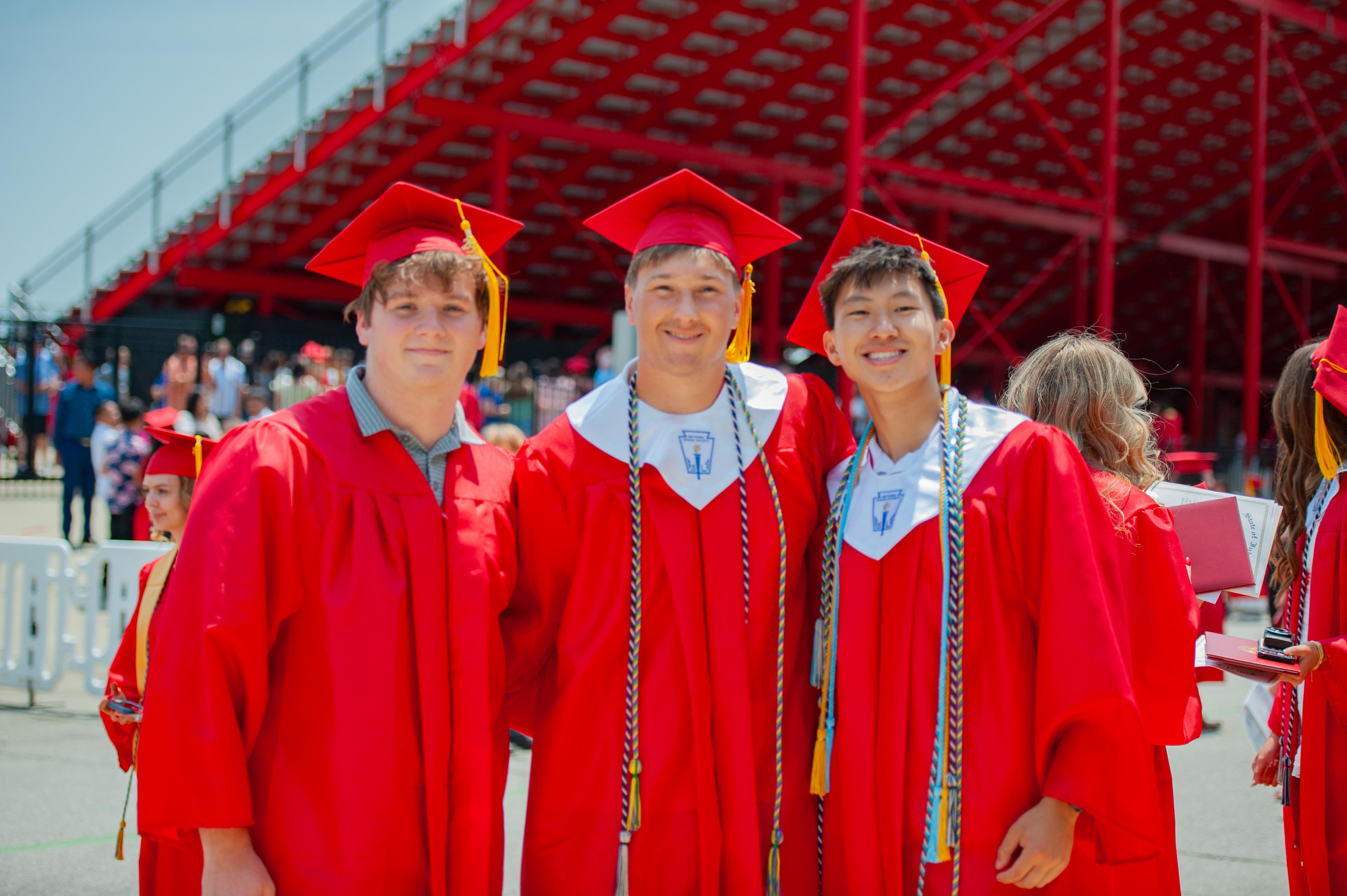 Three smiling graduates in red caps and gowns stand in the foreground with a blurred background of stadium seating and other graduates.