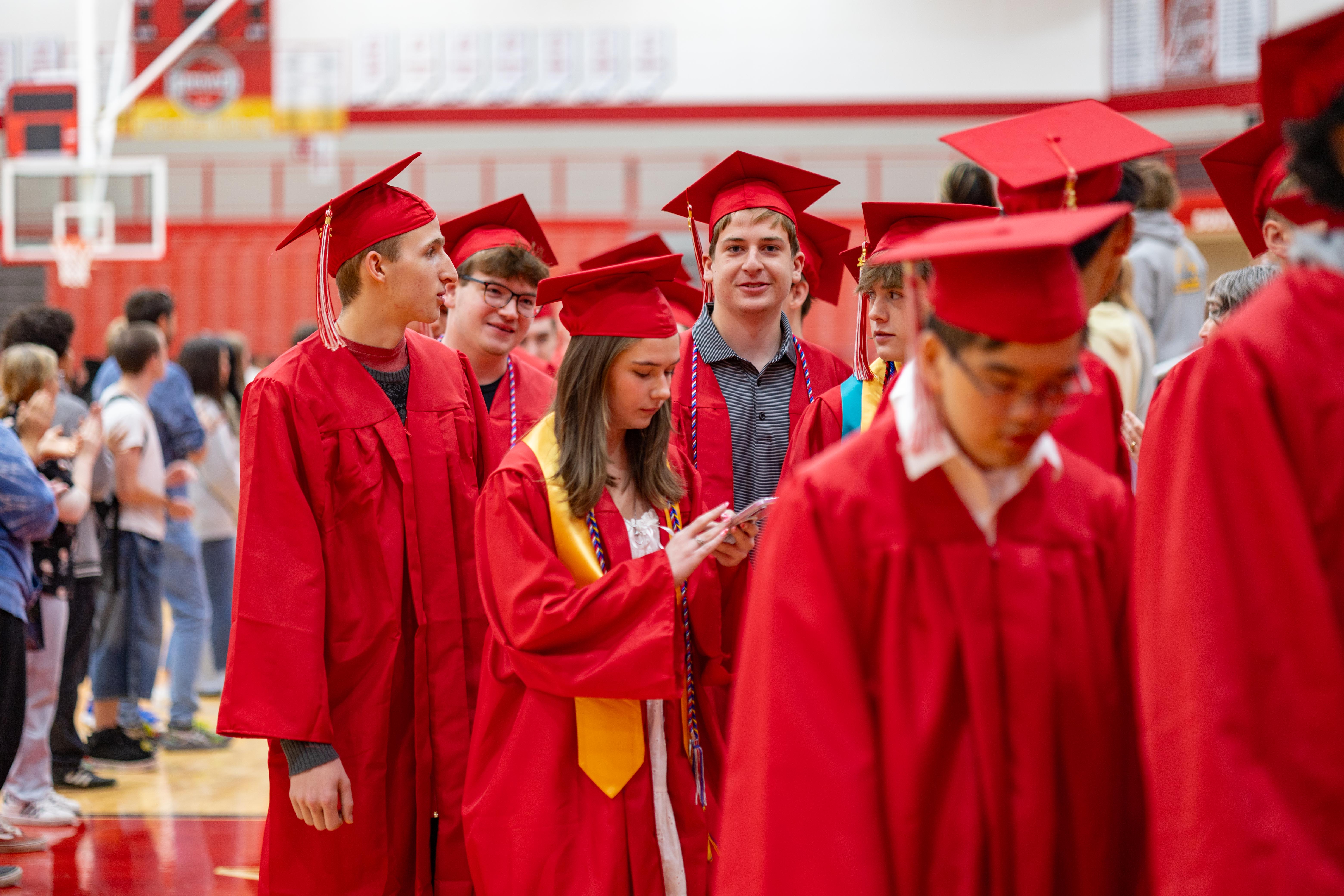 A group of graduates in red gowns and caps walk in a gymnasium with a basketball hoop visible in the background.