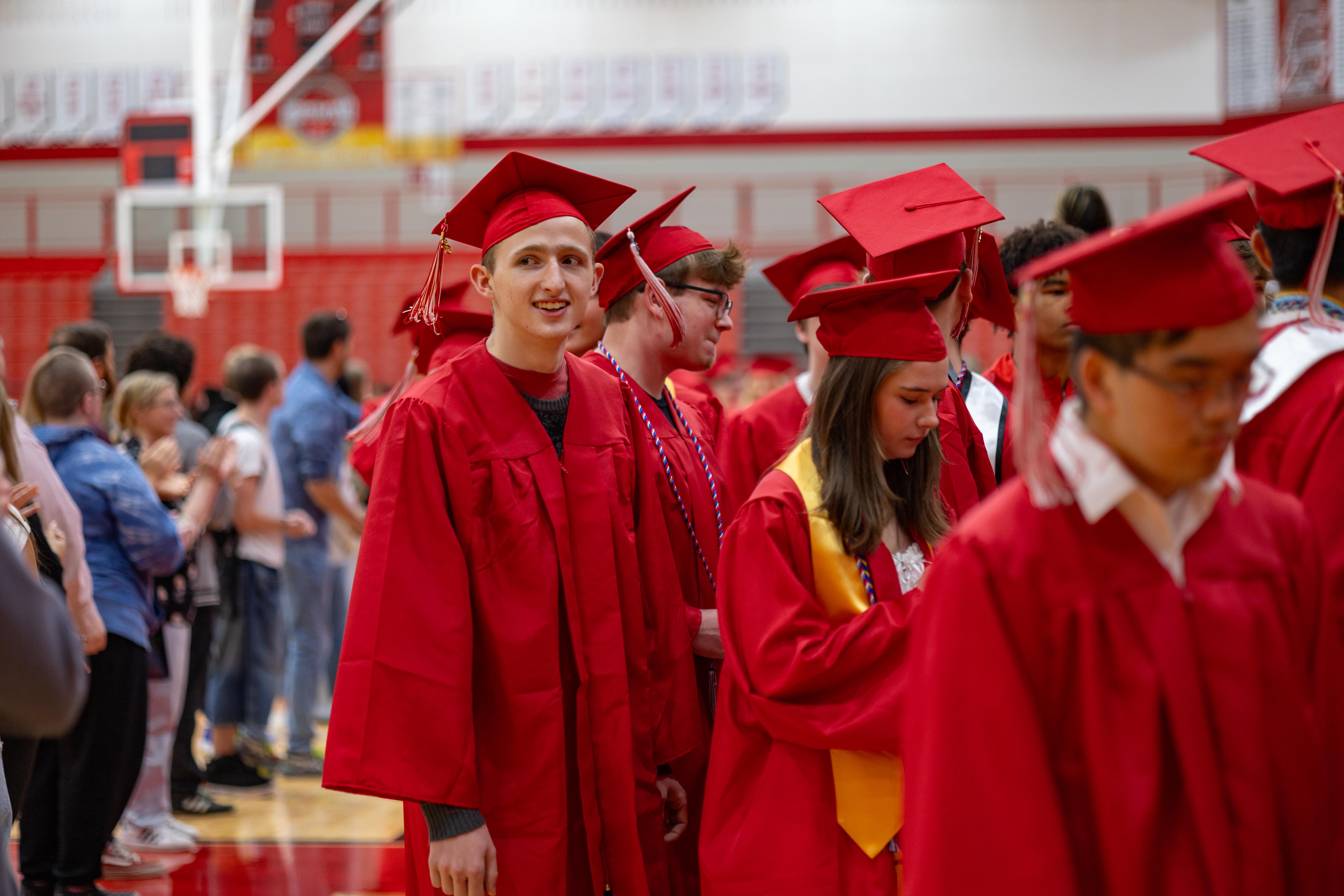 Graduates in red gowns and caps stand in a gymnasium with bleachers and a basketball hoop visible in the background.