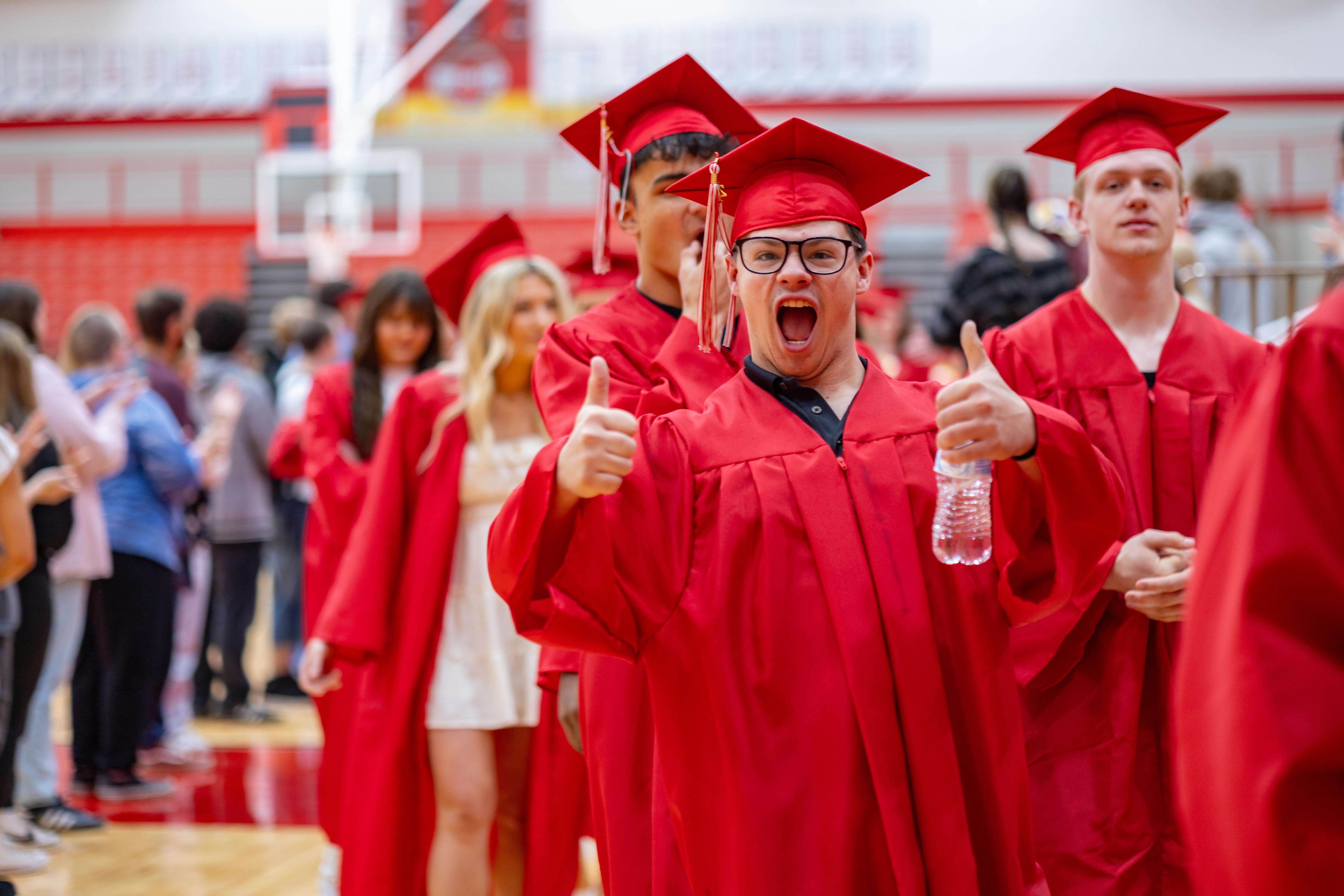 A group of excited graduates in red gowns and caps walk down a hallway, with a basketball hoop visible in the blurred background.