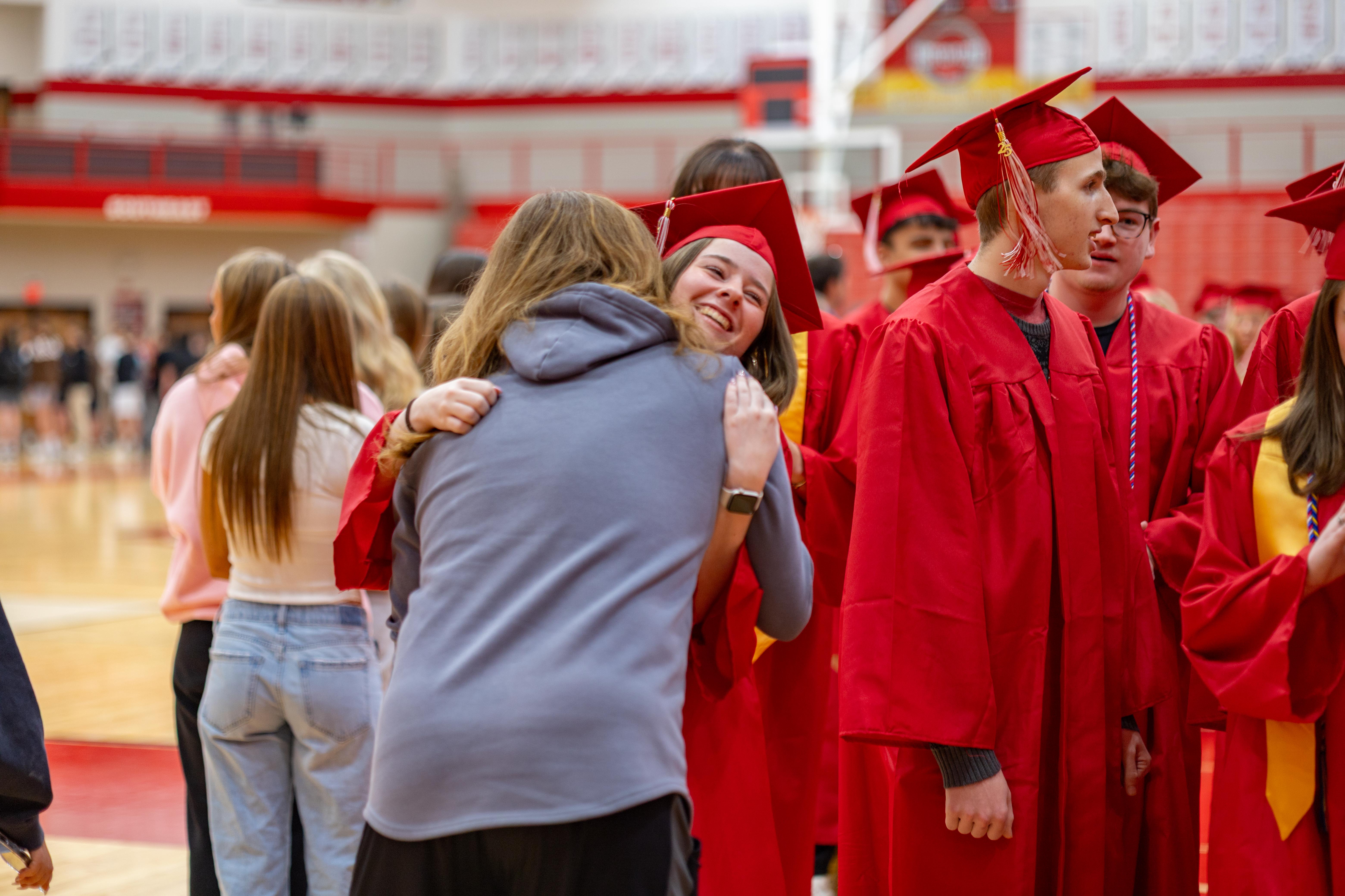 In the foreground, two people embrace, while in the background, a group of graduates in red gowns and caps stand in a gymnasium.