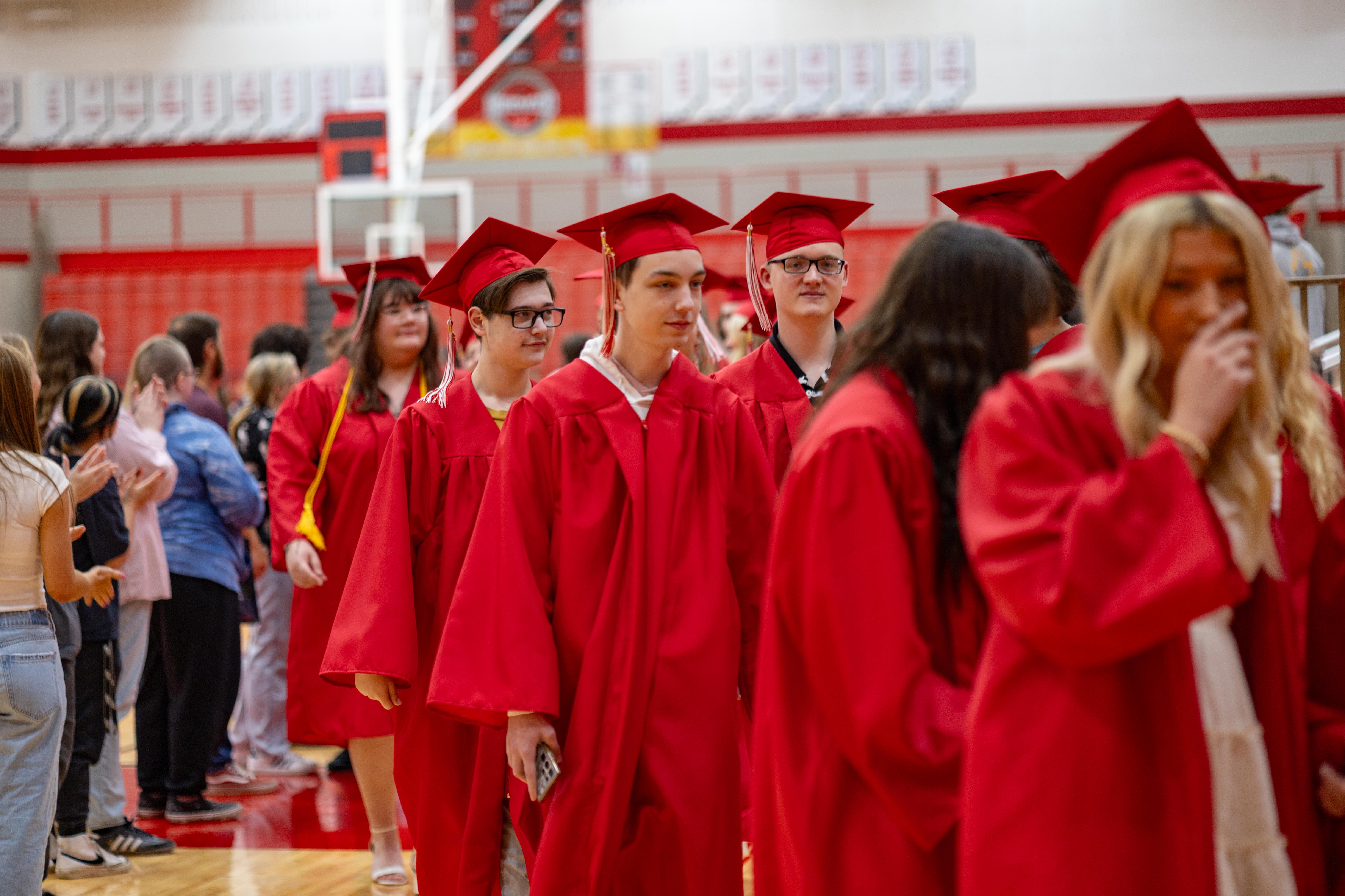 Graduates in red gowns and caps walk in a procession across a gym floor, with spectators visible in the blurred background.