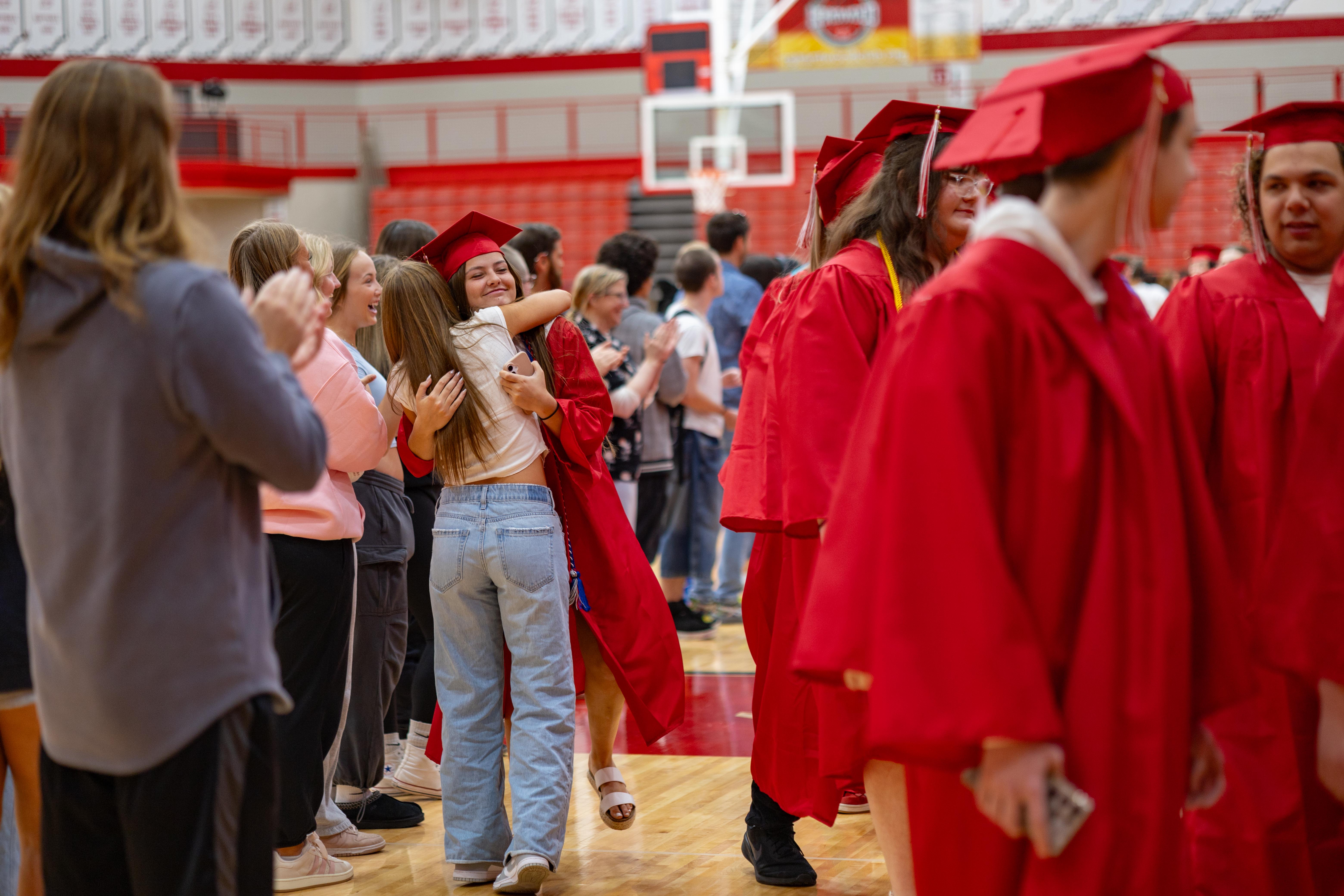 Graduates in red gowns hug in the foreground while others stand in the background of a gymnasium.