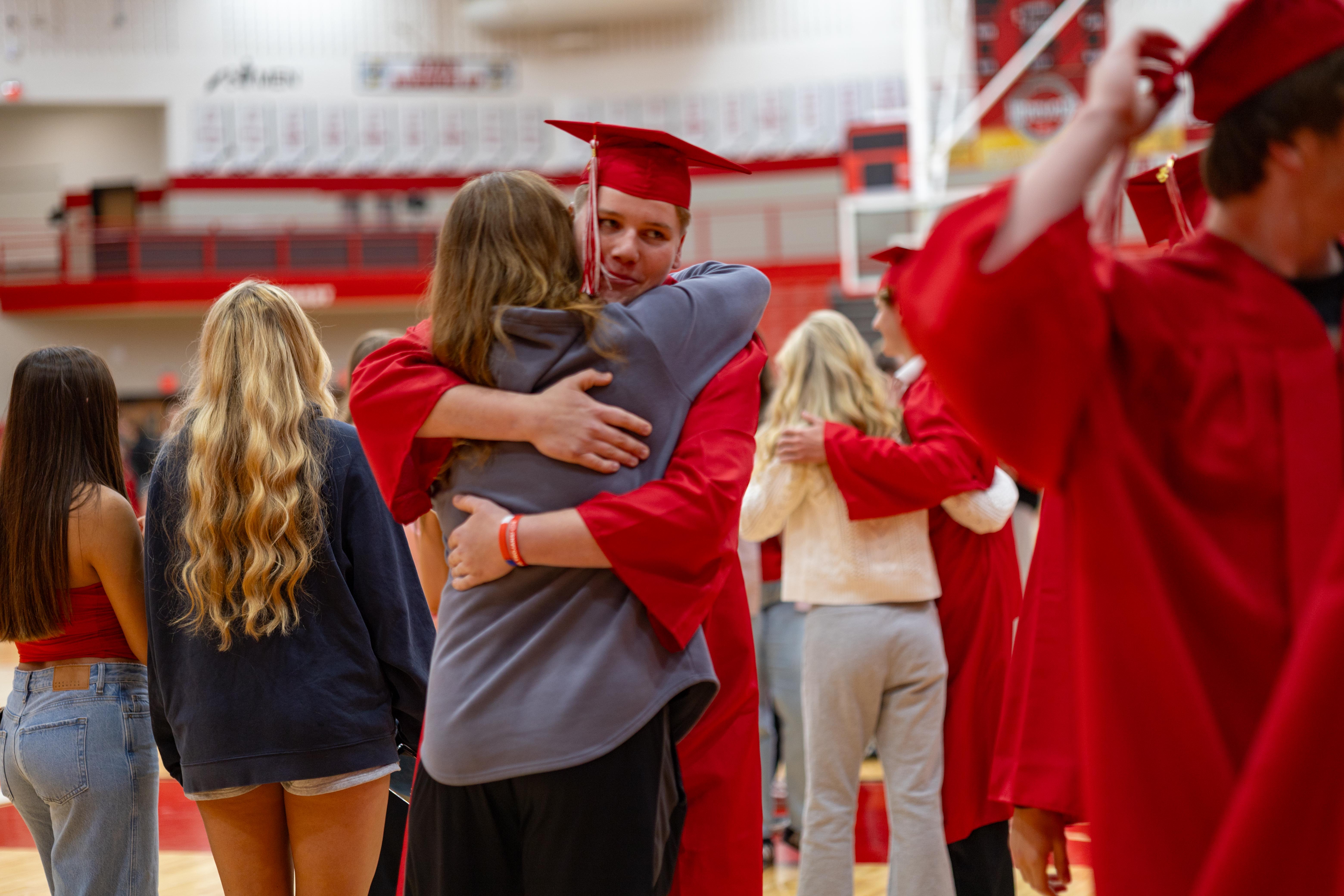 A graduating student in a red cap and gown hugs a friend in the foreground, while other graduates in red attire fill the background of a gymnasium.