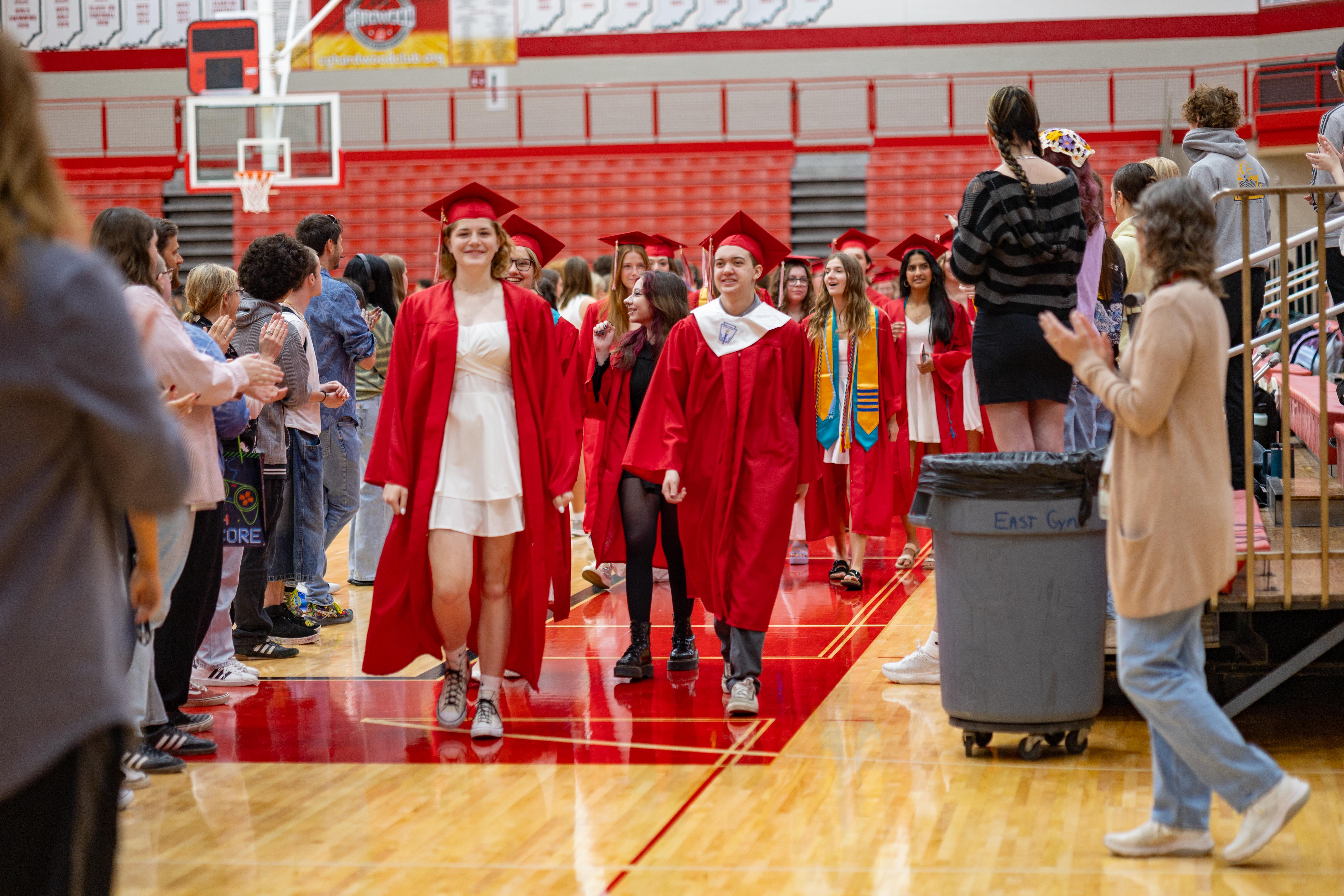 Graduates in red gowns and caps walk down a gymnasium floor lined with spectators, with bleachers and a basketball hoop visible in the background.
