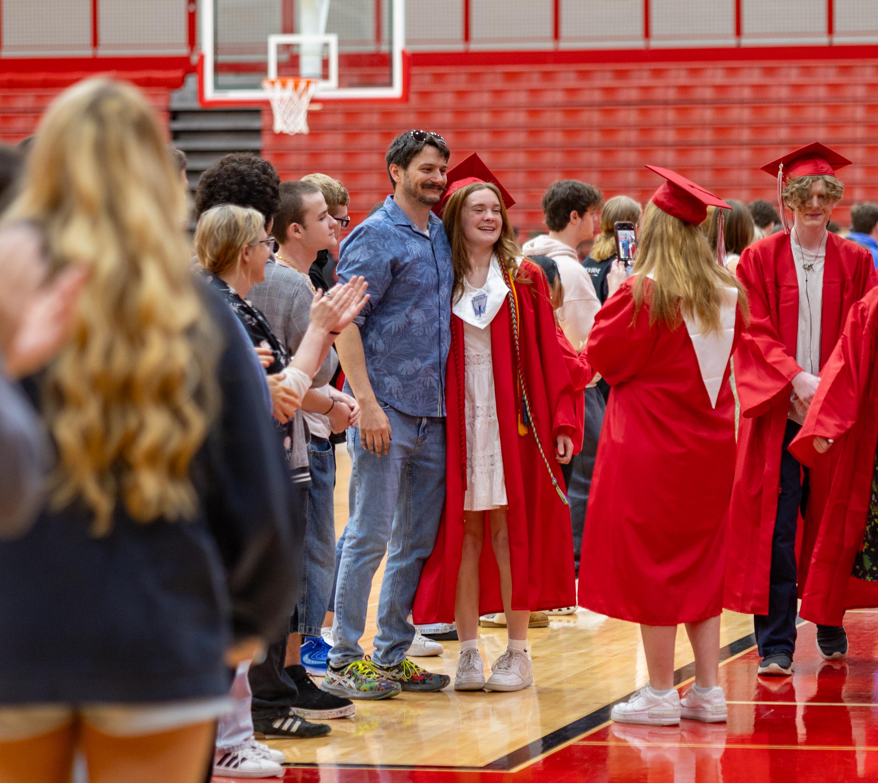 Graduates in red gowns and caps walk past cheering onlookers in a gymnasium with red bleachers and a basketball hoop in the background.