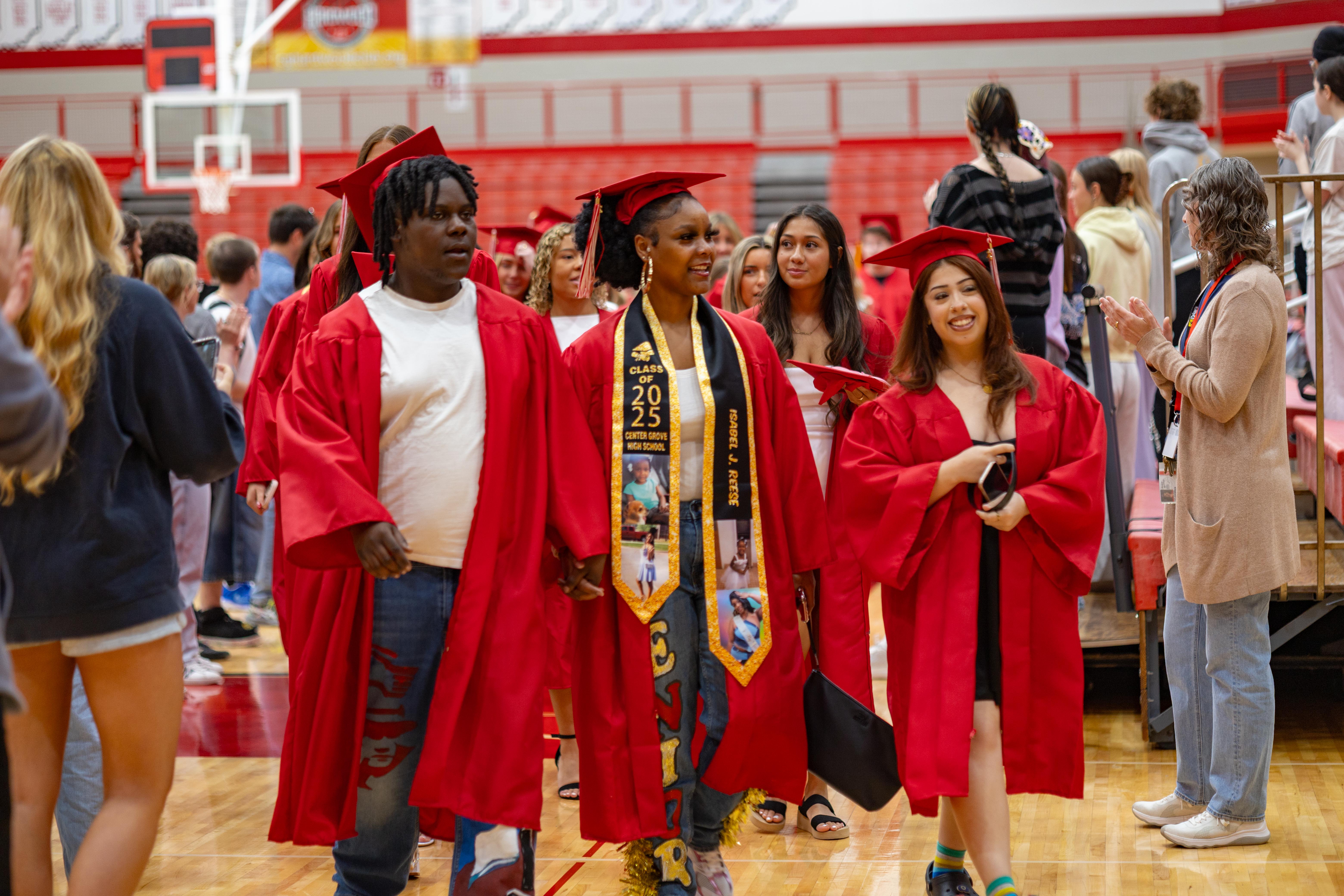Graduates in red gowns walk across a gymnasium floor with spectators in the background.