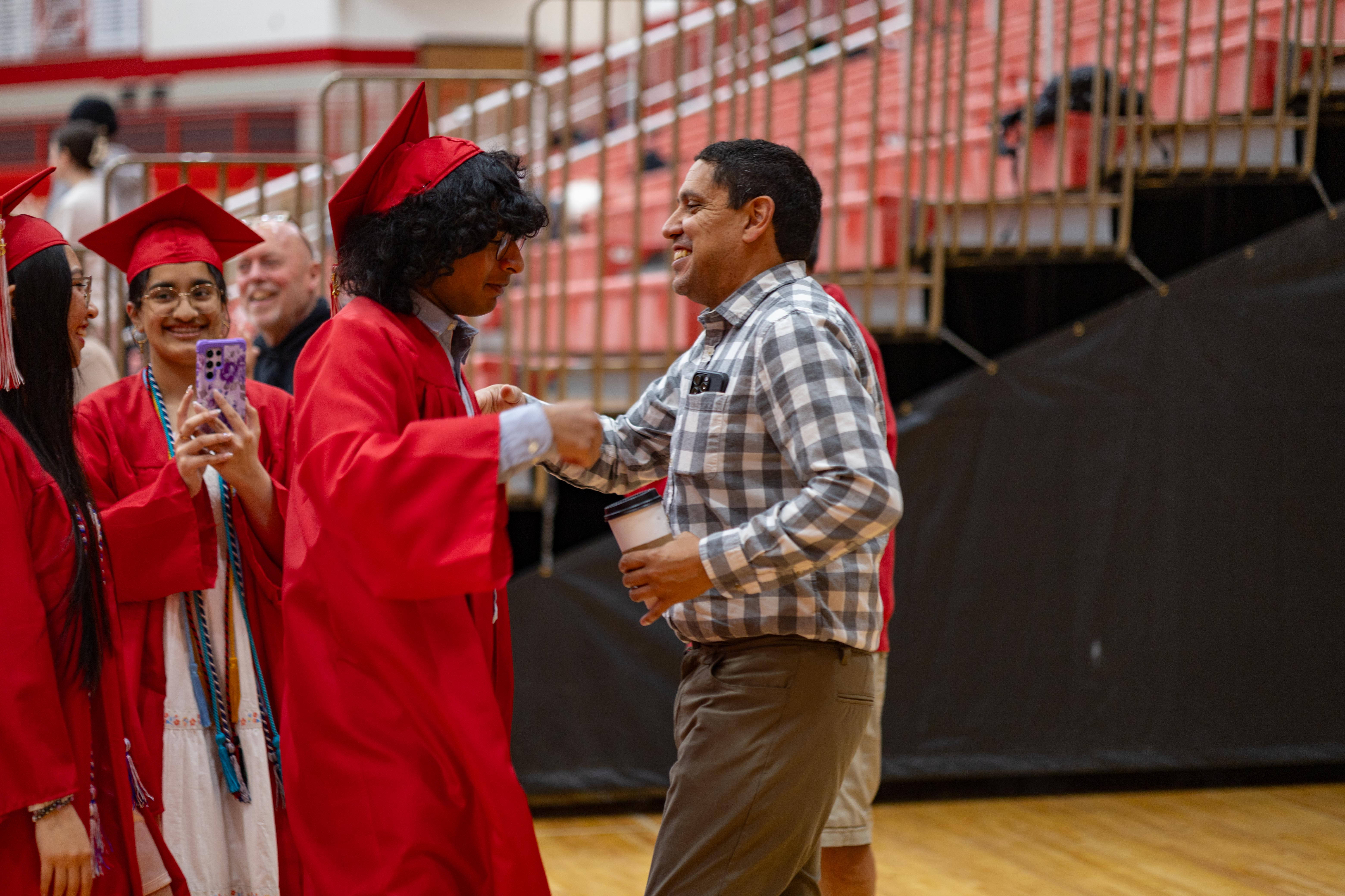 A graduate in a red cap and gown shakes hands with a man in a plaid shirt, while other graduates and bleachers fill the background.