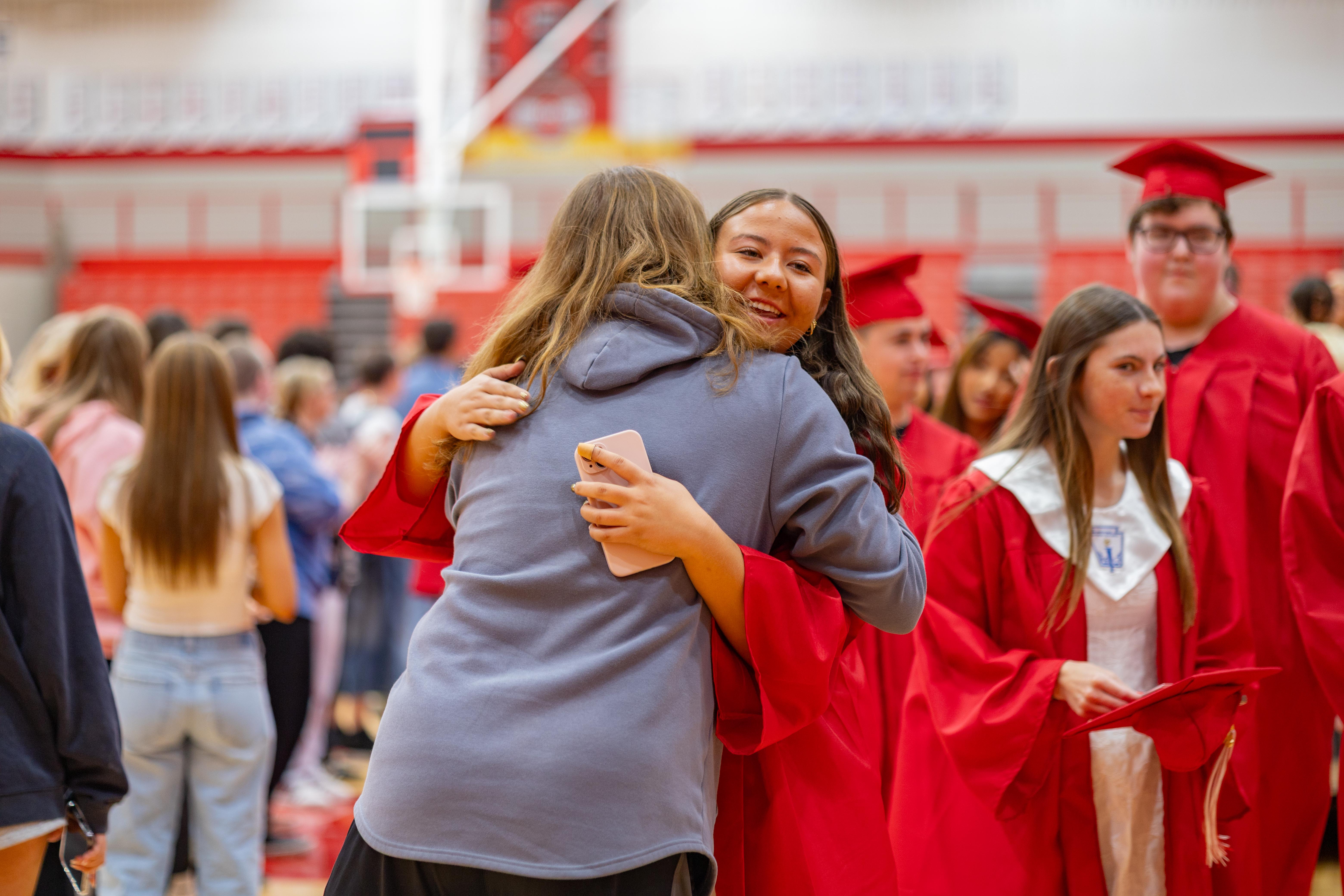 Two young women in graduation gowns embrace in the foreground, while other graduates and spectators fill the blurred background of a gymnasium.