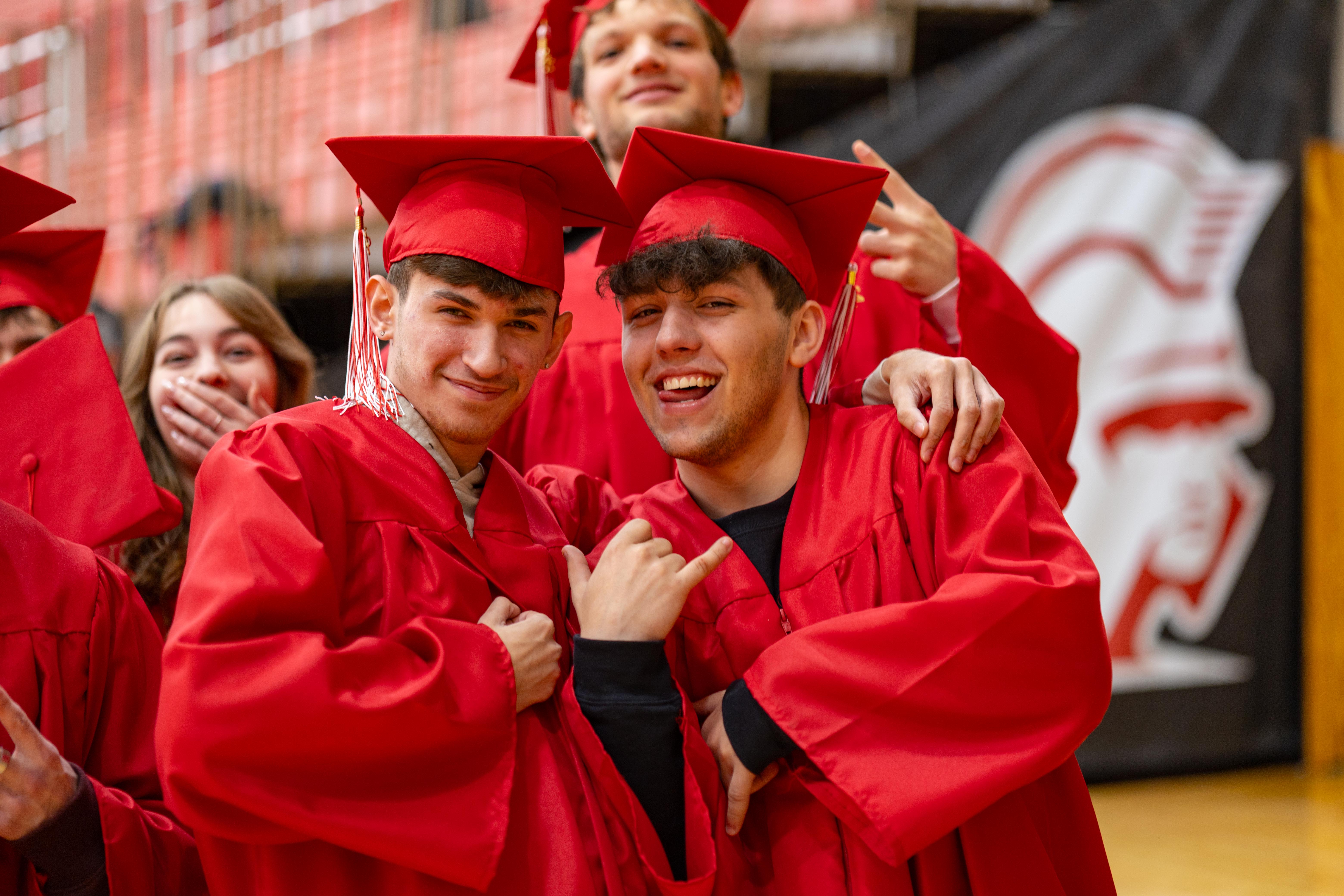 Two smiling young men in red graduation gowns and caps pose in the foreground, with other graduates and a large school mascot banner visible in the blurred background.