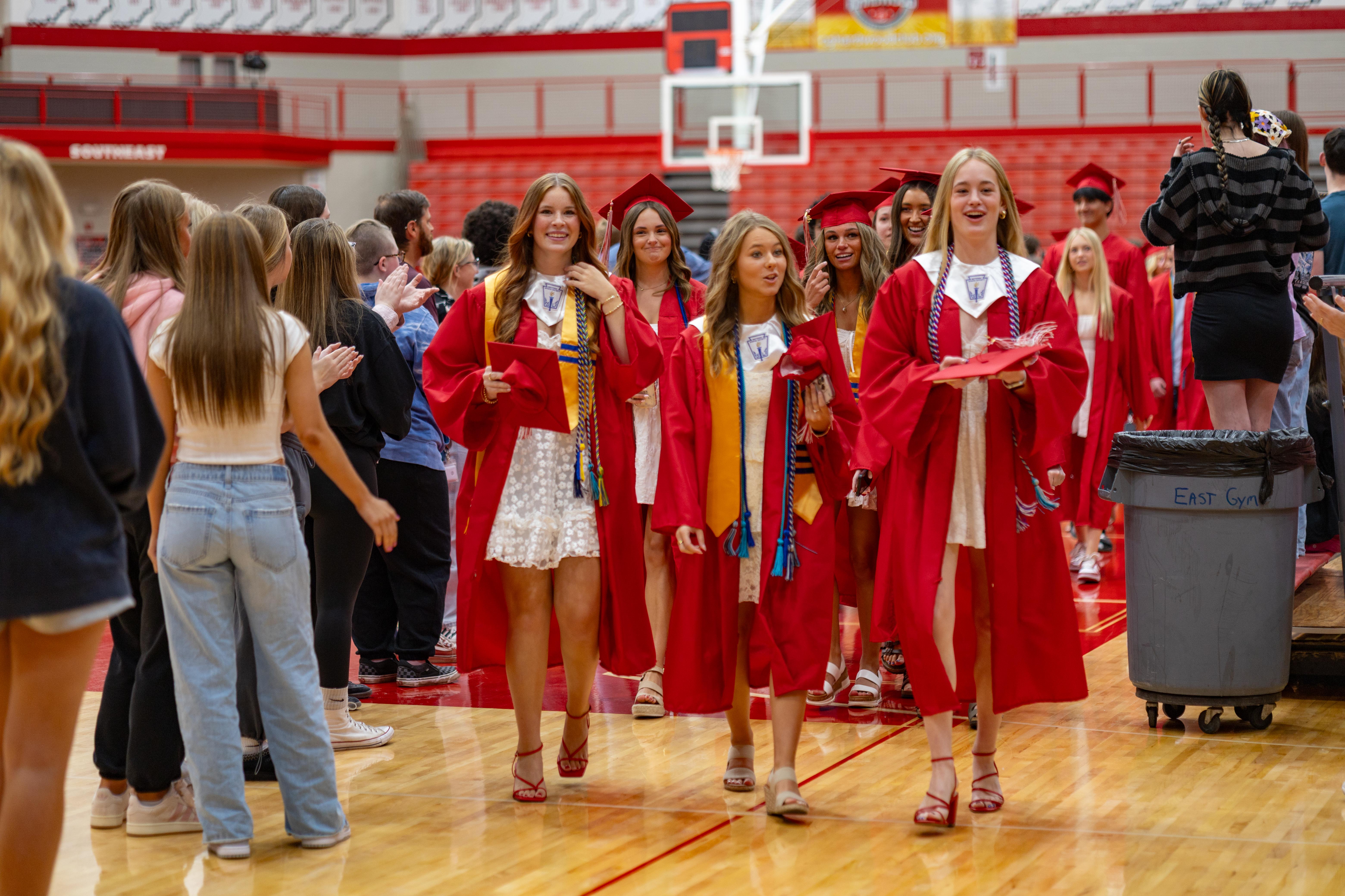Graduating students in red gowns walk across a polished gym floor towards the camera, while a crowd of onlookers watches from the bleachers in the background.