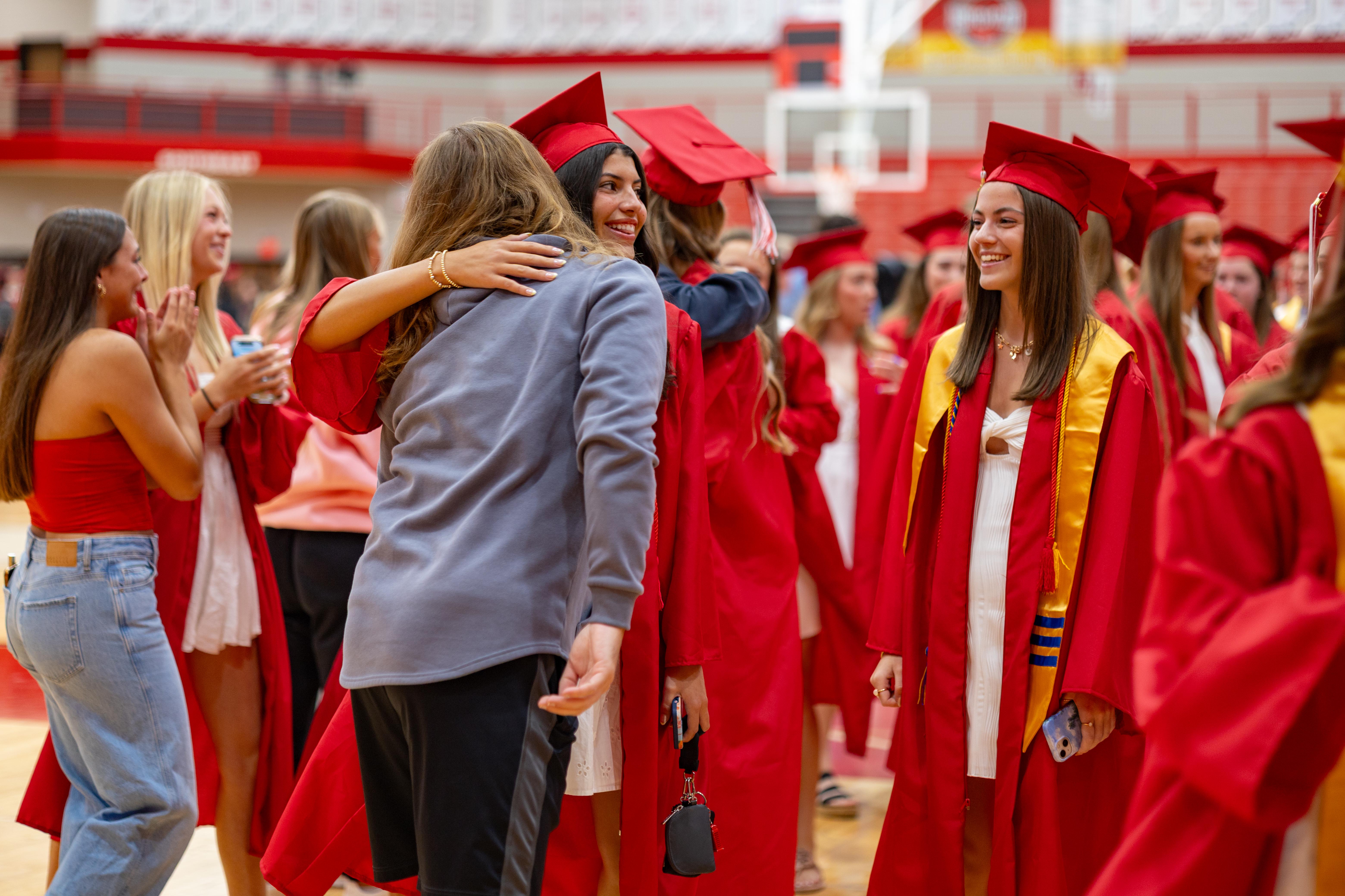 In the foreground, graduates in red gowns and caps embrace and smile, while the background shows a gymnasium with bleachers.