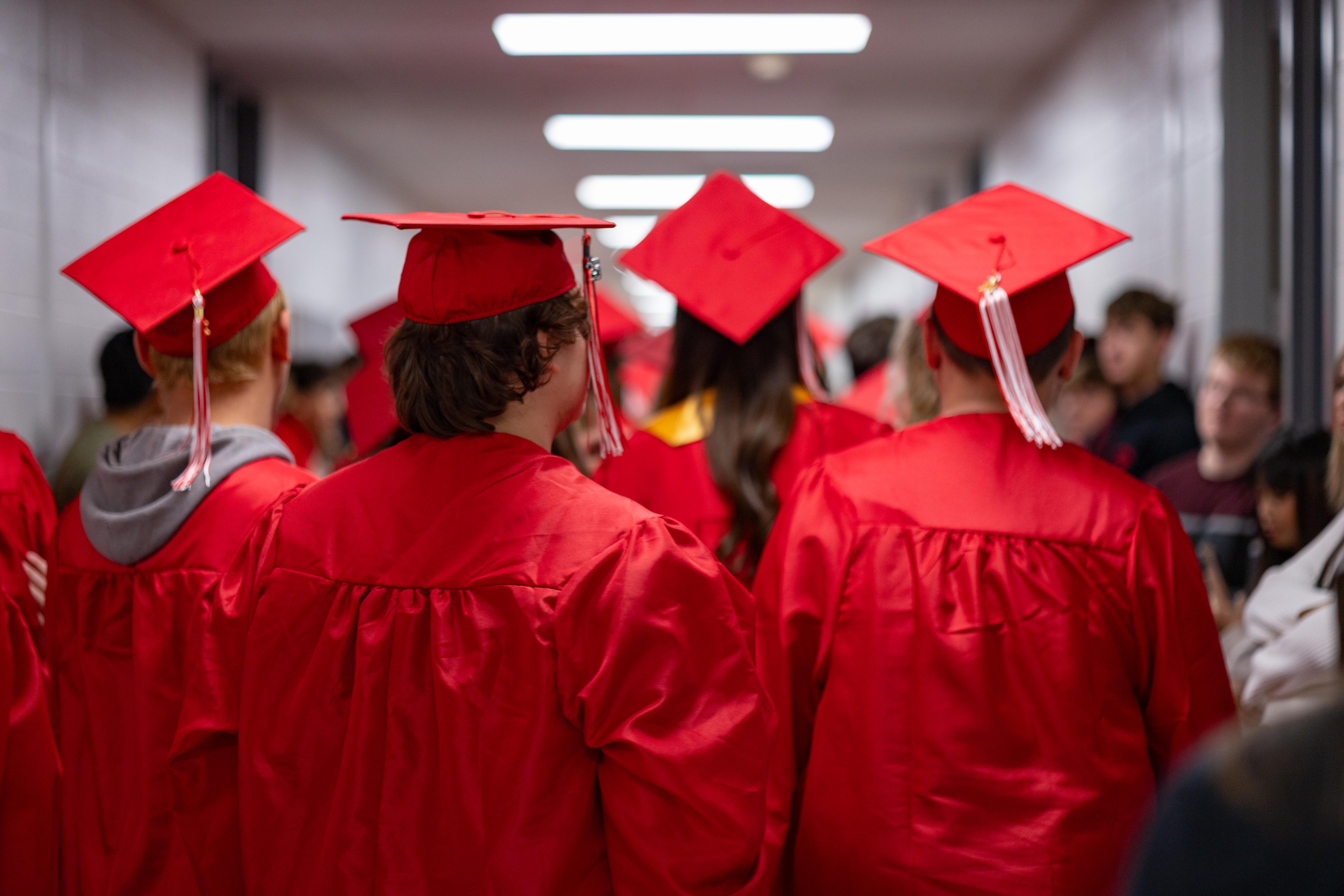 Graduates in red gowns and caps walk down a hallway with blurred figures in the background.
