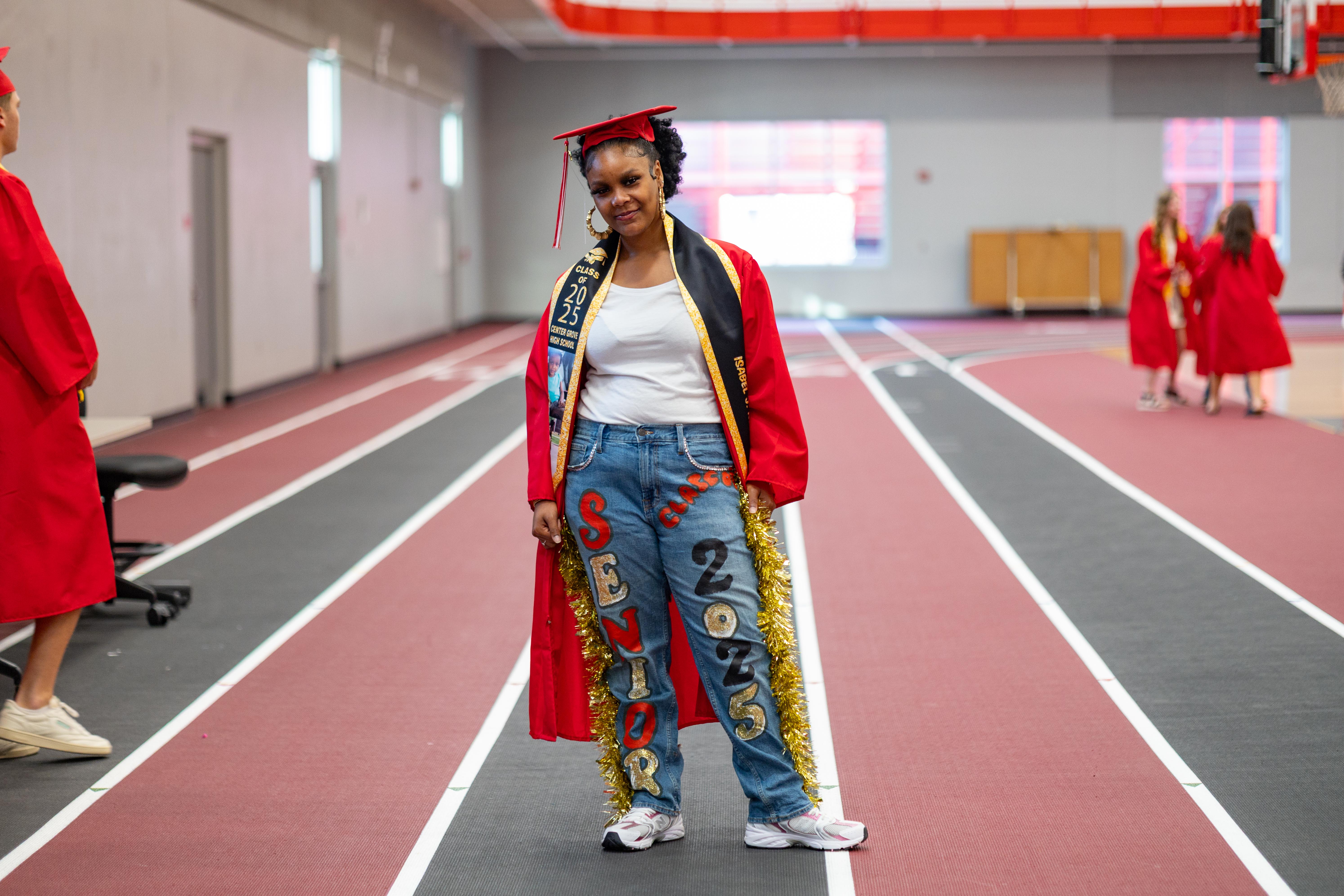 A graduate in a red cap and gown with decorated jeans stands on a red track, while other graduates in similar attire are visible in the background of an indoor athletic facility.