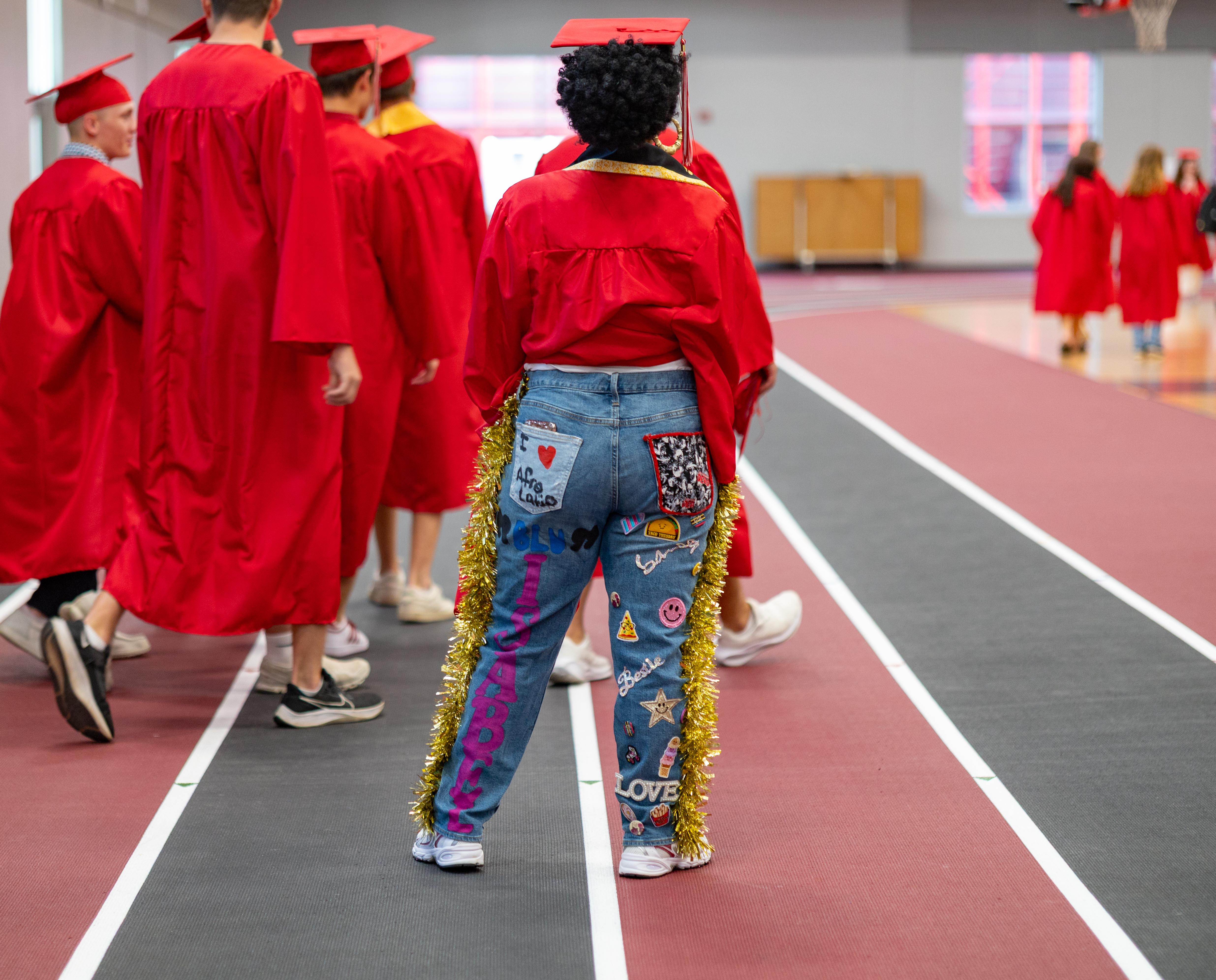 In the foreground, a graduate in decorated jeans and a red cap and gown walks away from the camera, while in the background, other graduates in red gowns are seen in a gymnasium.
