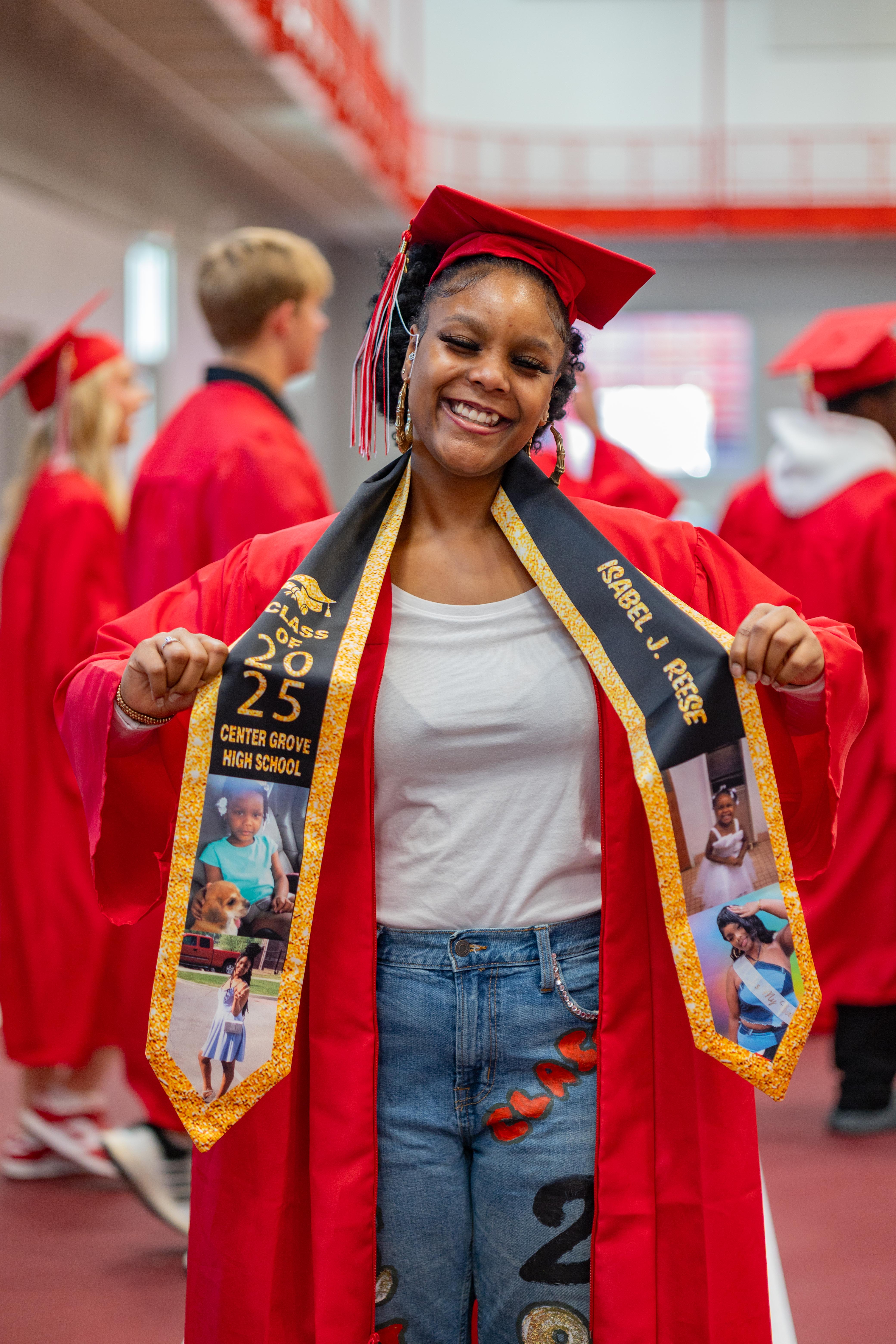 A smiling graduate in a red cap and gown, adorned with a personalized sash featuring photos and text, stands in a hallway with other graduates in the background.