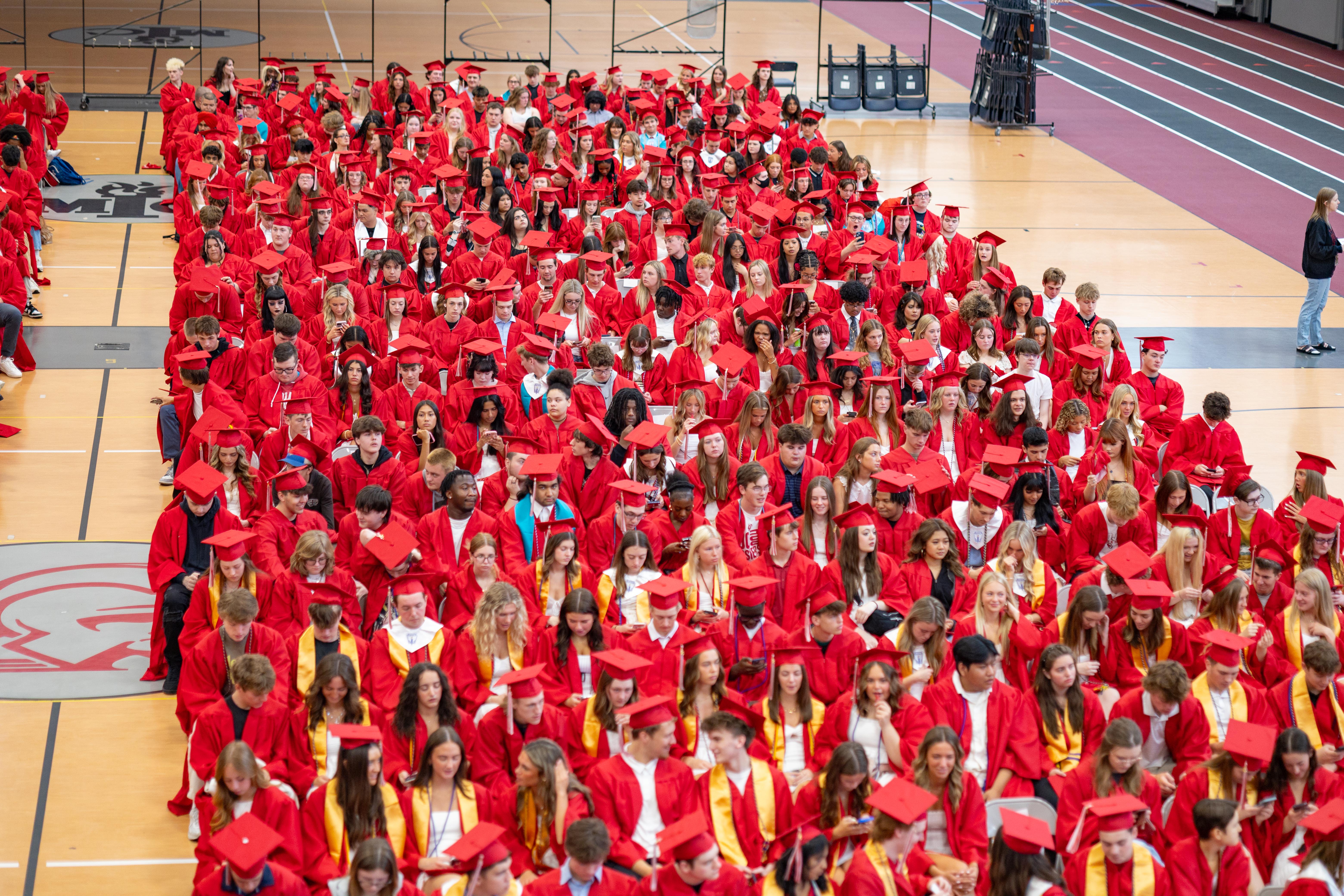 A large group of graduates in red gowns fill the foreground, while the background shows a gymnasium with bleachers and a track.