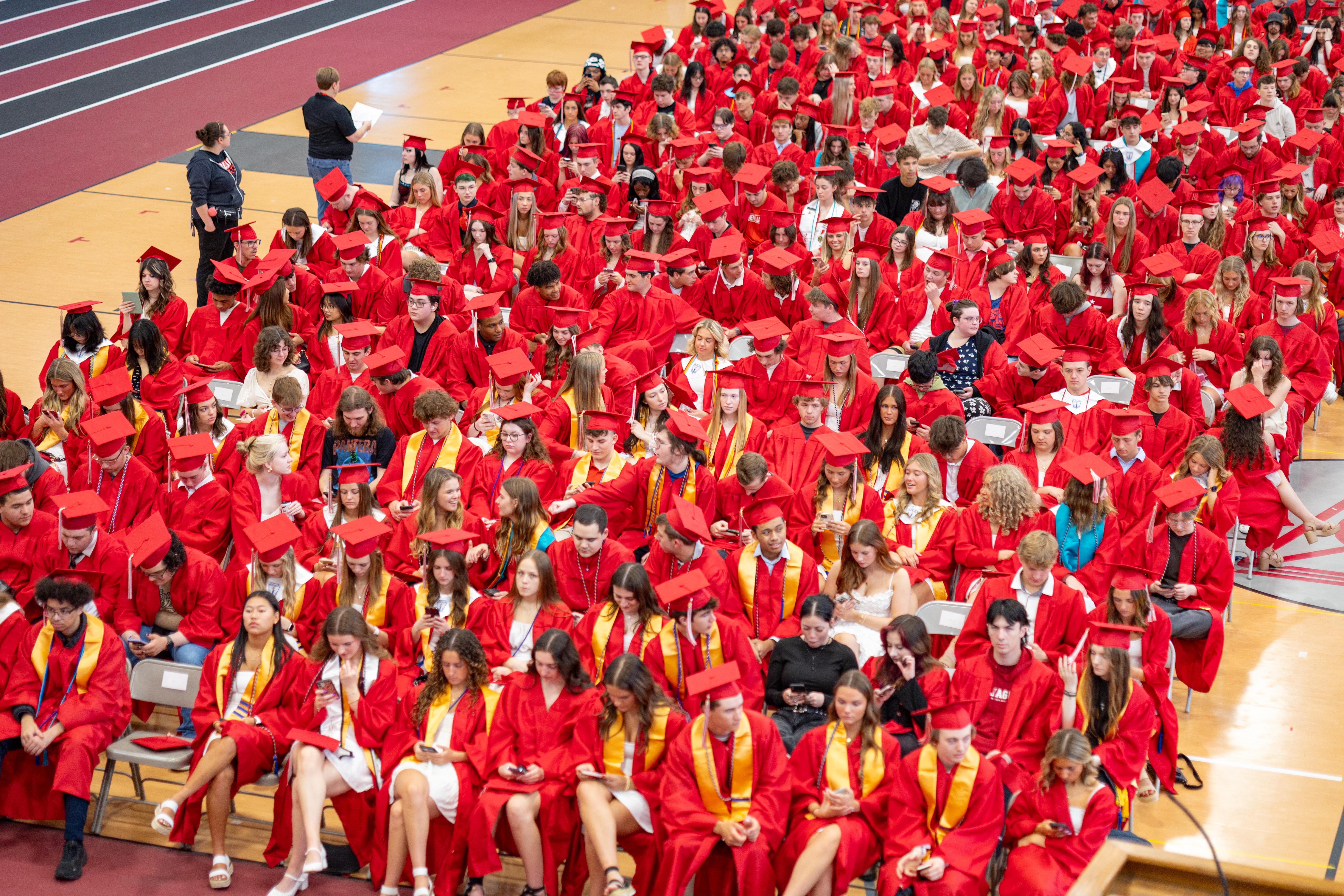 A large crowd of graduates in red gowns and caps fill a gymnasium, with a few people standing and addressing them in the foreground.