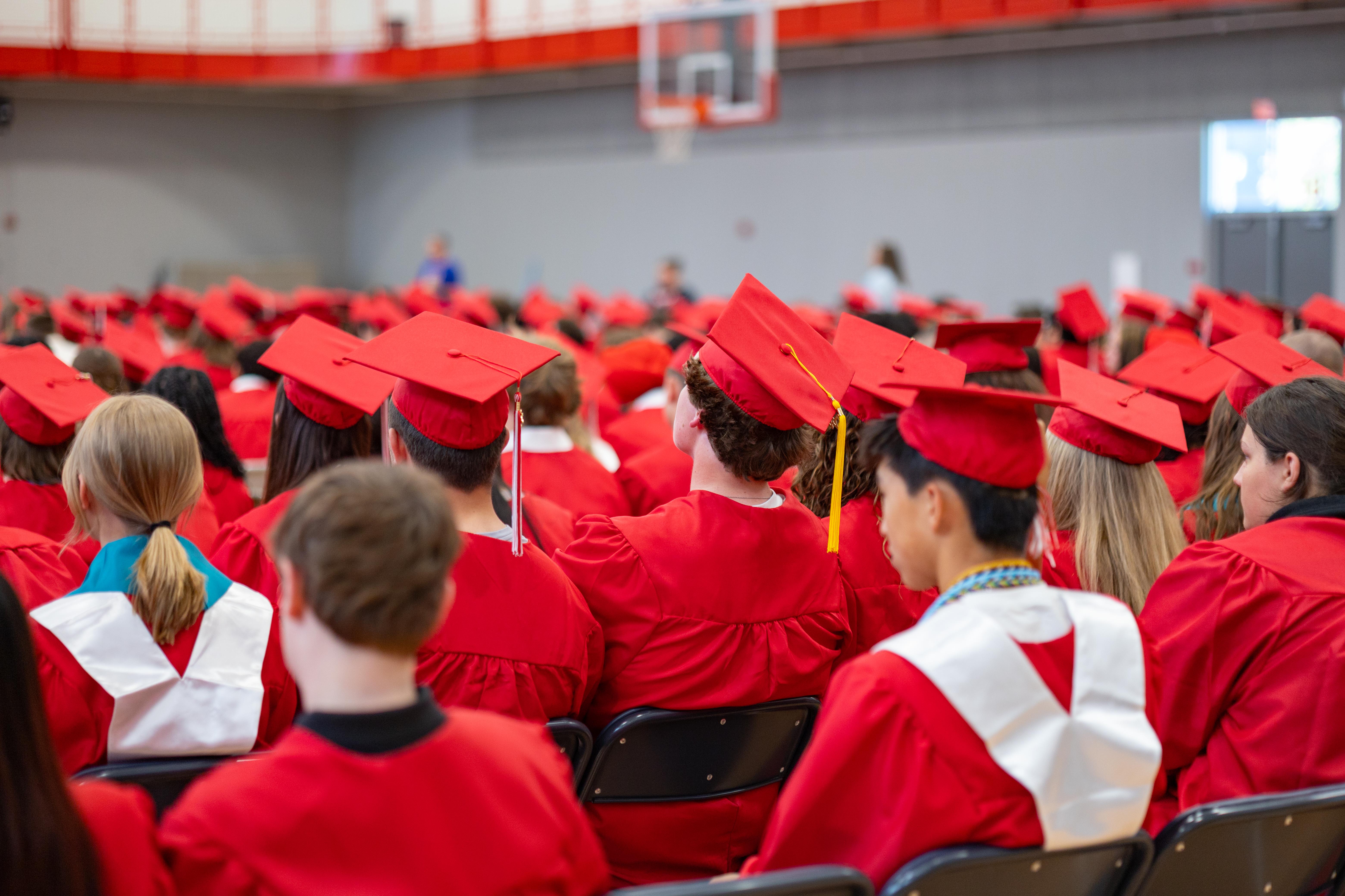 Graduates in red caps and gowns fill the foreground, while a blurred gymnasium with spectators forms the background.