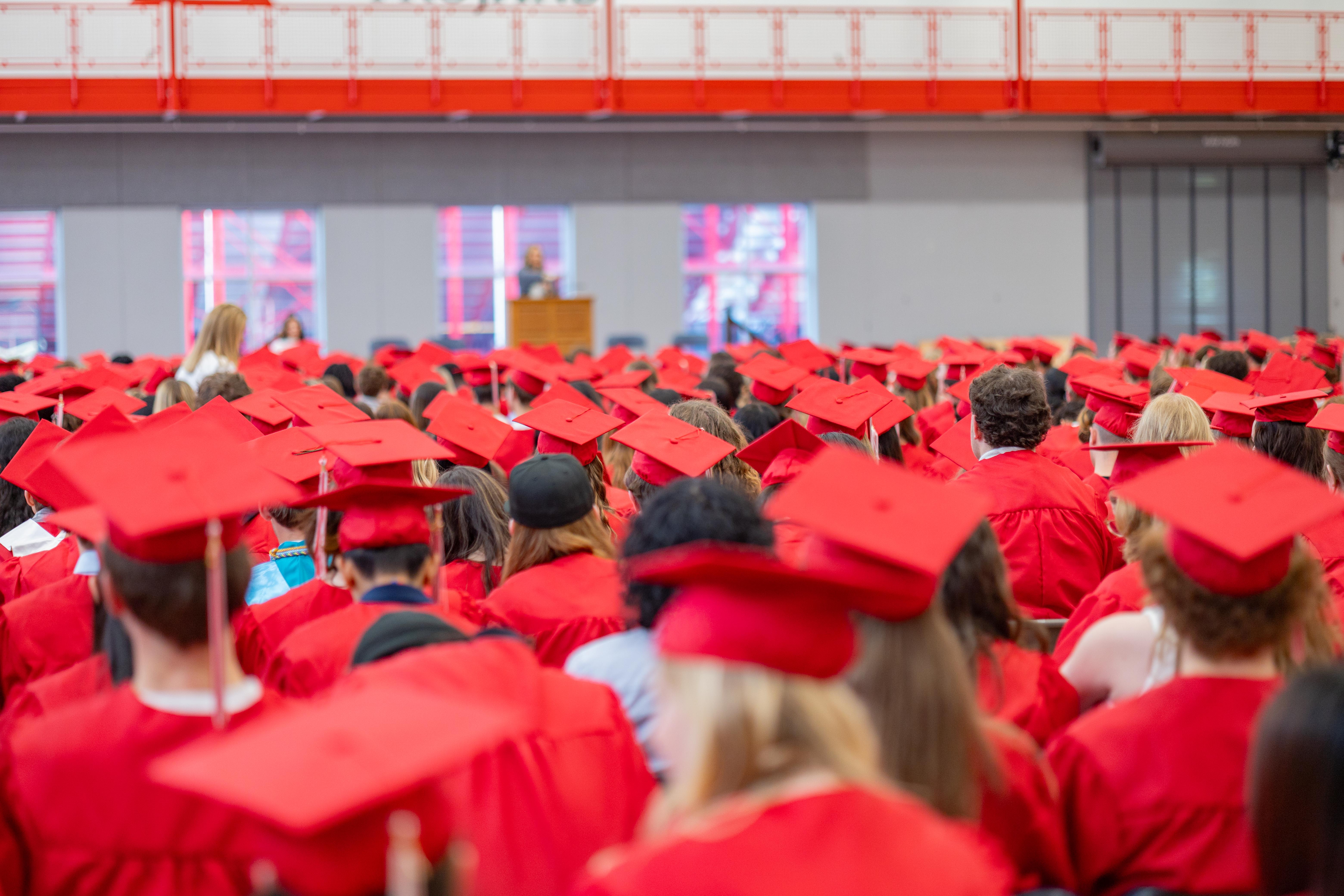 A crowd of graduates in red caps and gowns fills the foreground, while a speaker stands at a podium in the blurred background.
