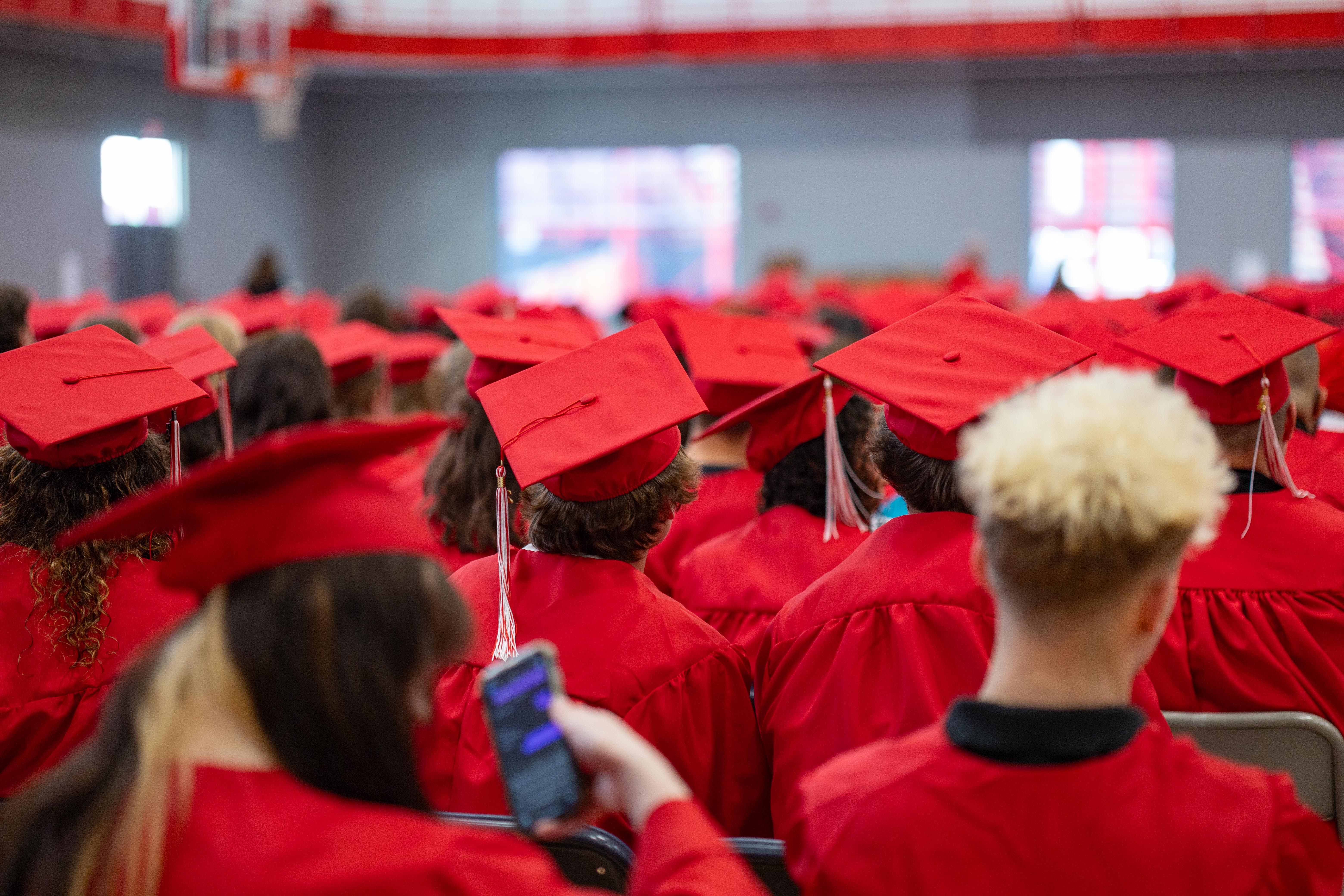 Graduates in red gowns and caps fill the foreground, while a blurred gymnasium with red accents forms the background.