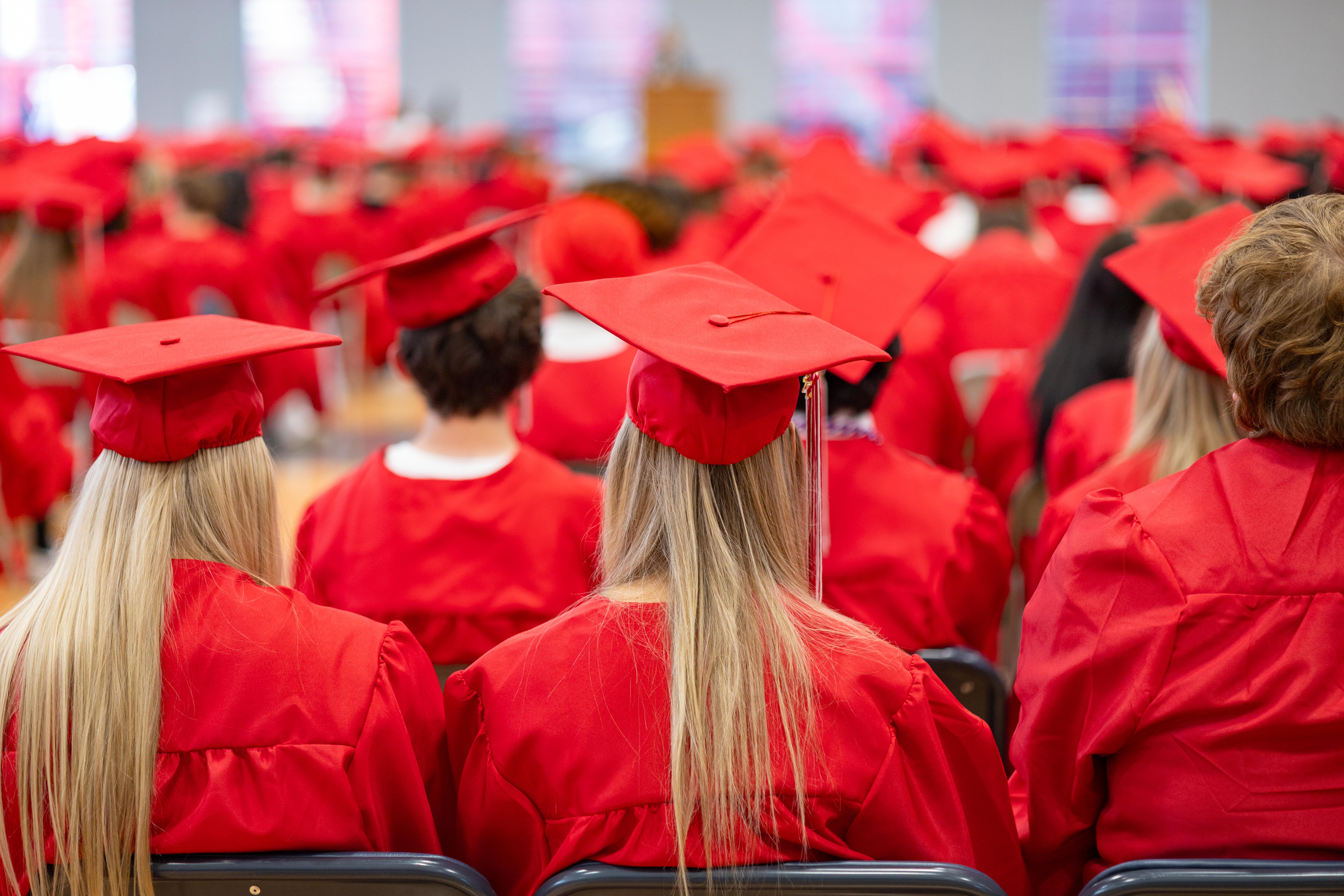 Graduates in red gowns and caps are seated in the foreground, facing a blurred background of more graduates and a speaker at a podium.