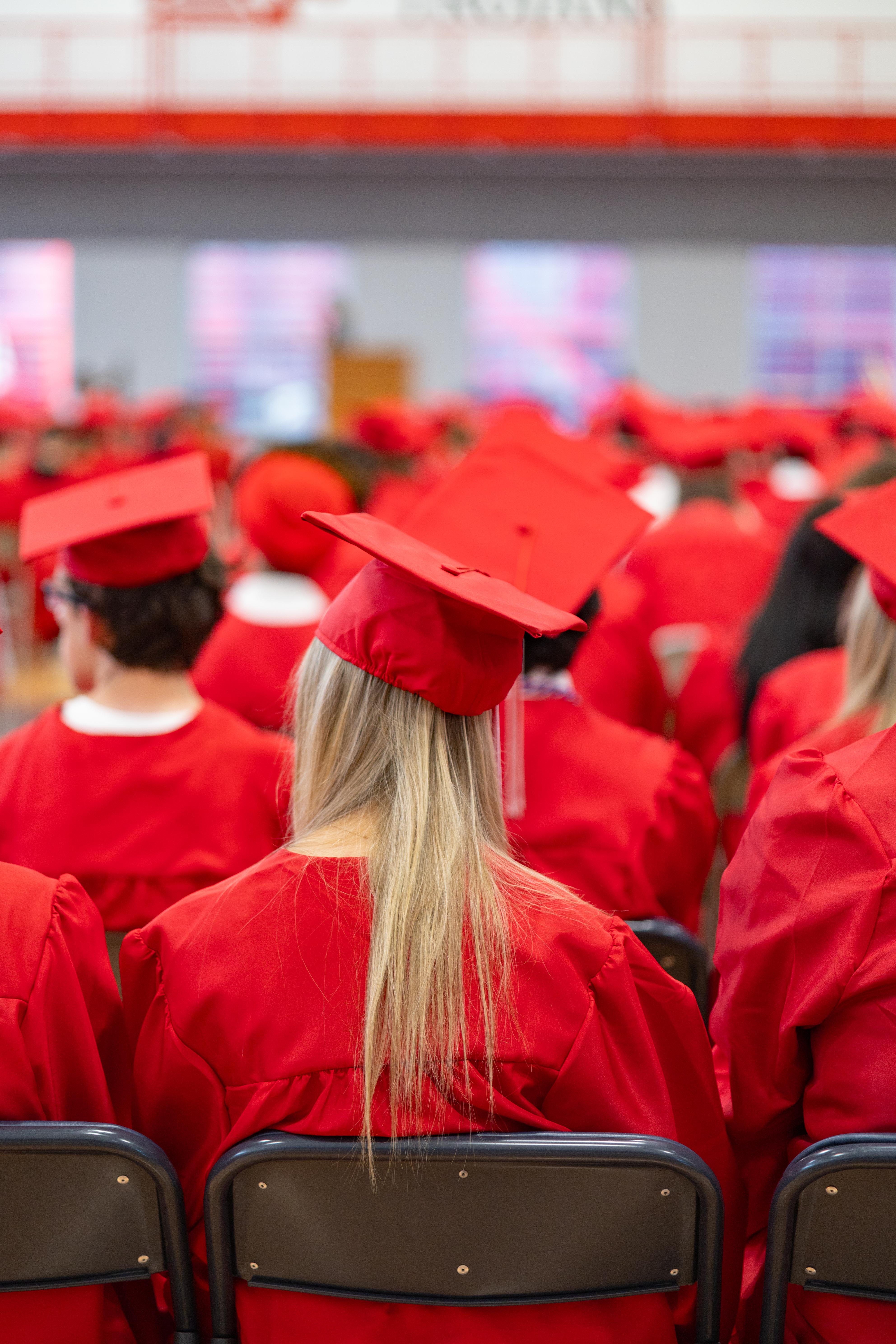 A blonde-haired graduate in a red cap and gown sits in the foreground, facing away from the camera, while a blurred background shows rows of other graduates and a stage.