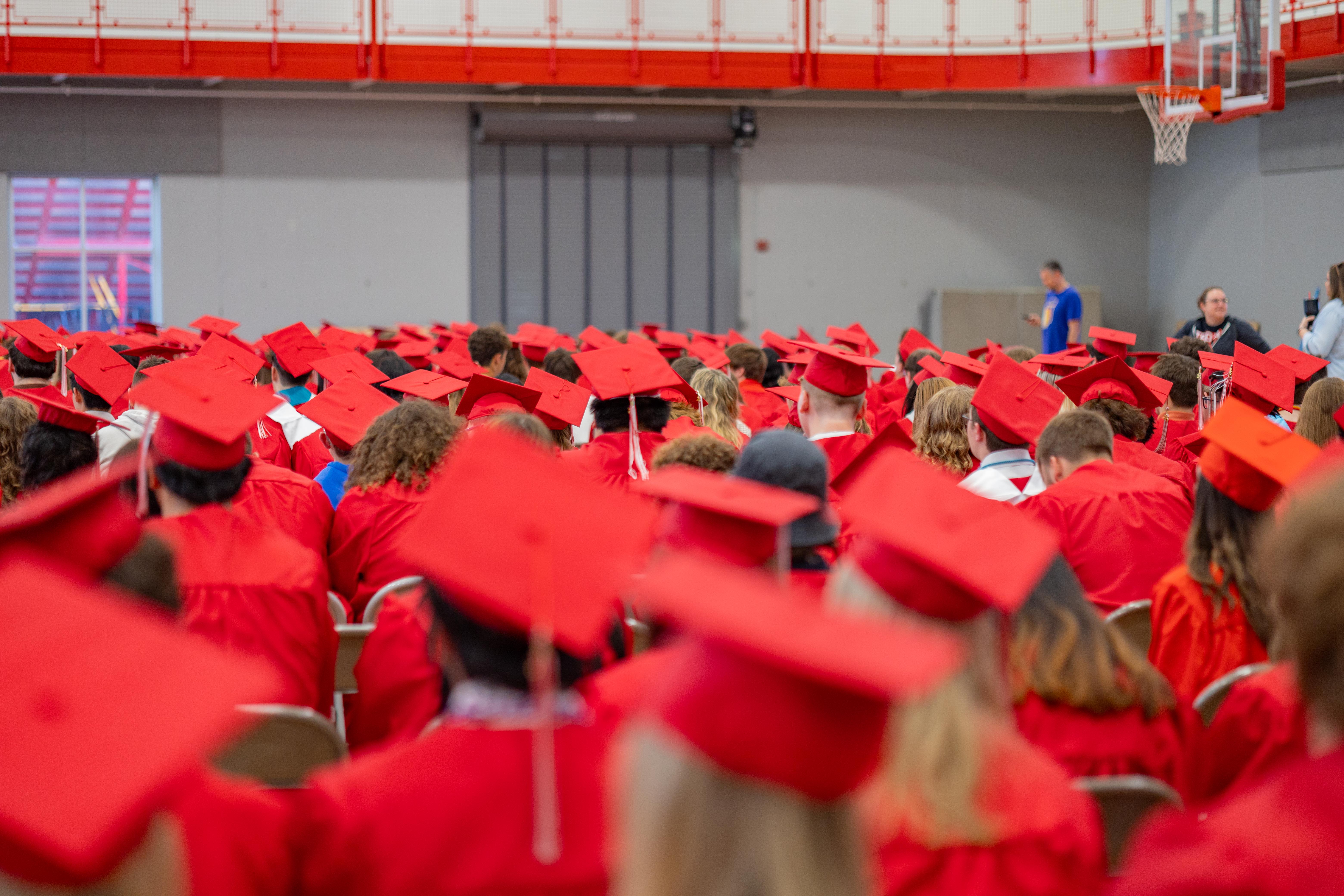 A crowd of graduates in red gowns and caps fills the foreground of a gymnasium, with a basketball hoop visible in the blurred background.