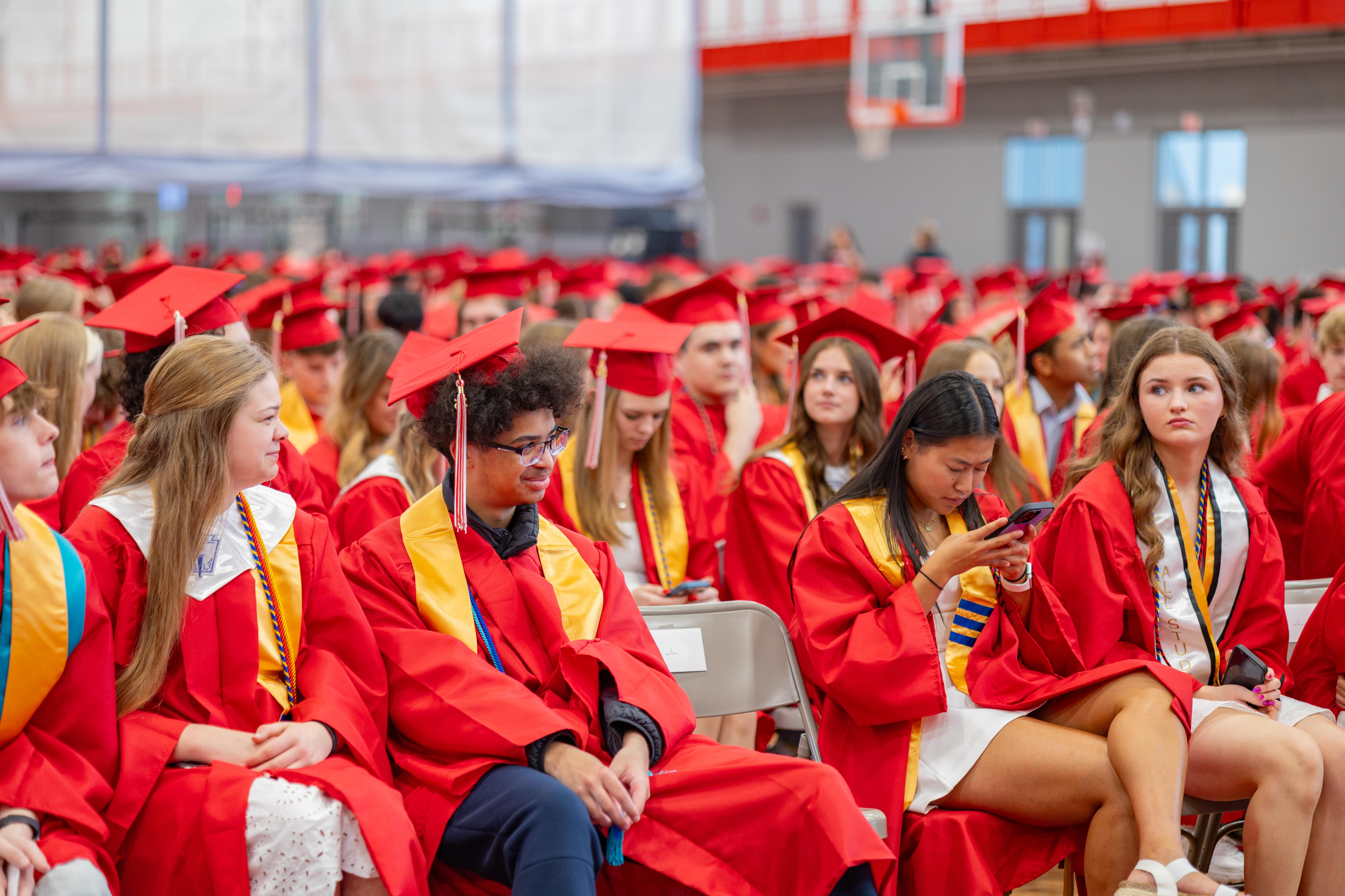 Graduates in red gowns and caps sit in chairs in a large gymnasium, with a basketball hoop visible in the blurred background.