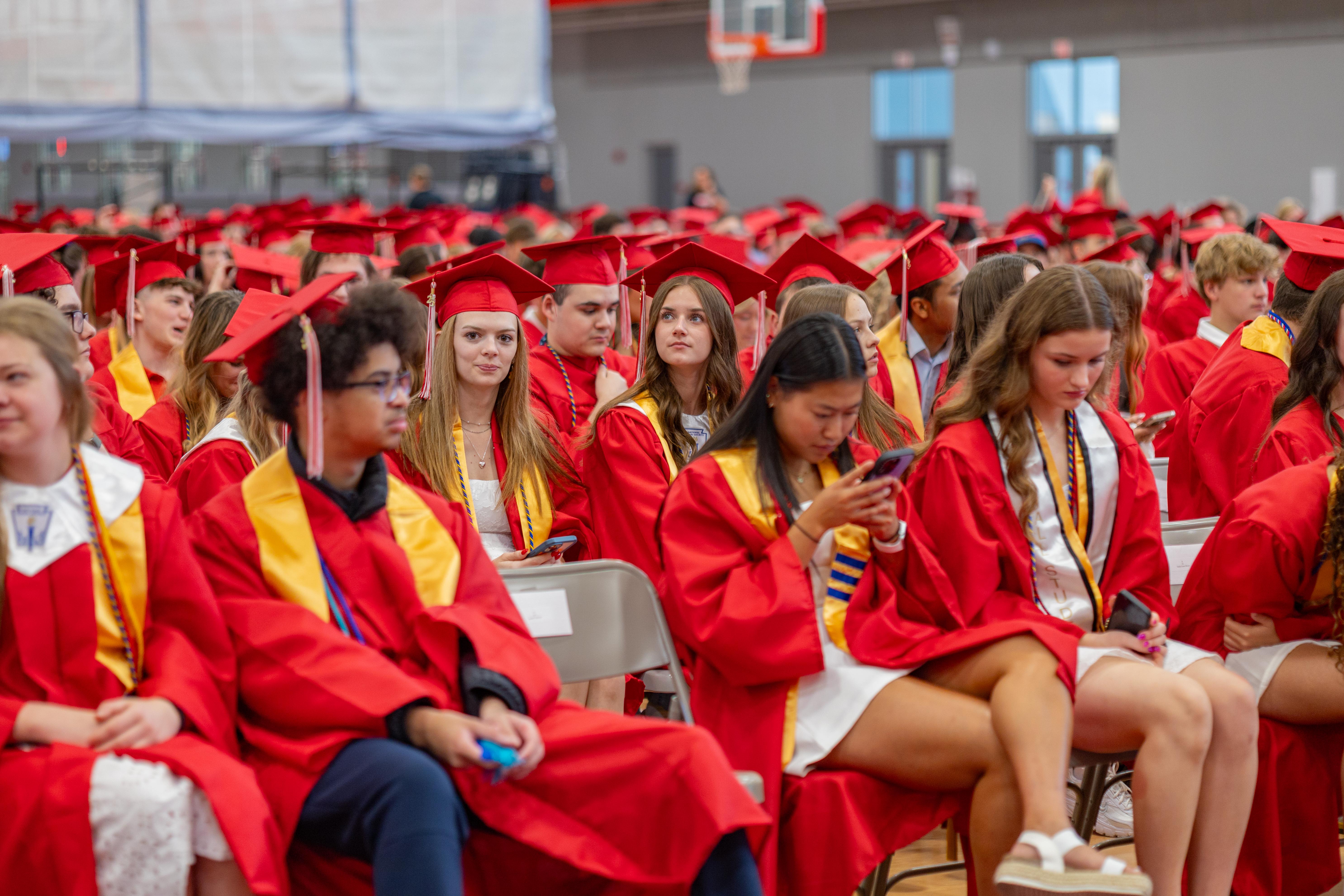 Graduates in red gowns and caps sit in the foreground, while a blurred gymnasium with more graduates and spectators fills the background.