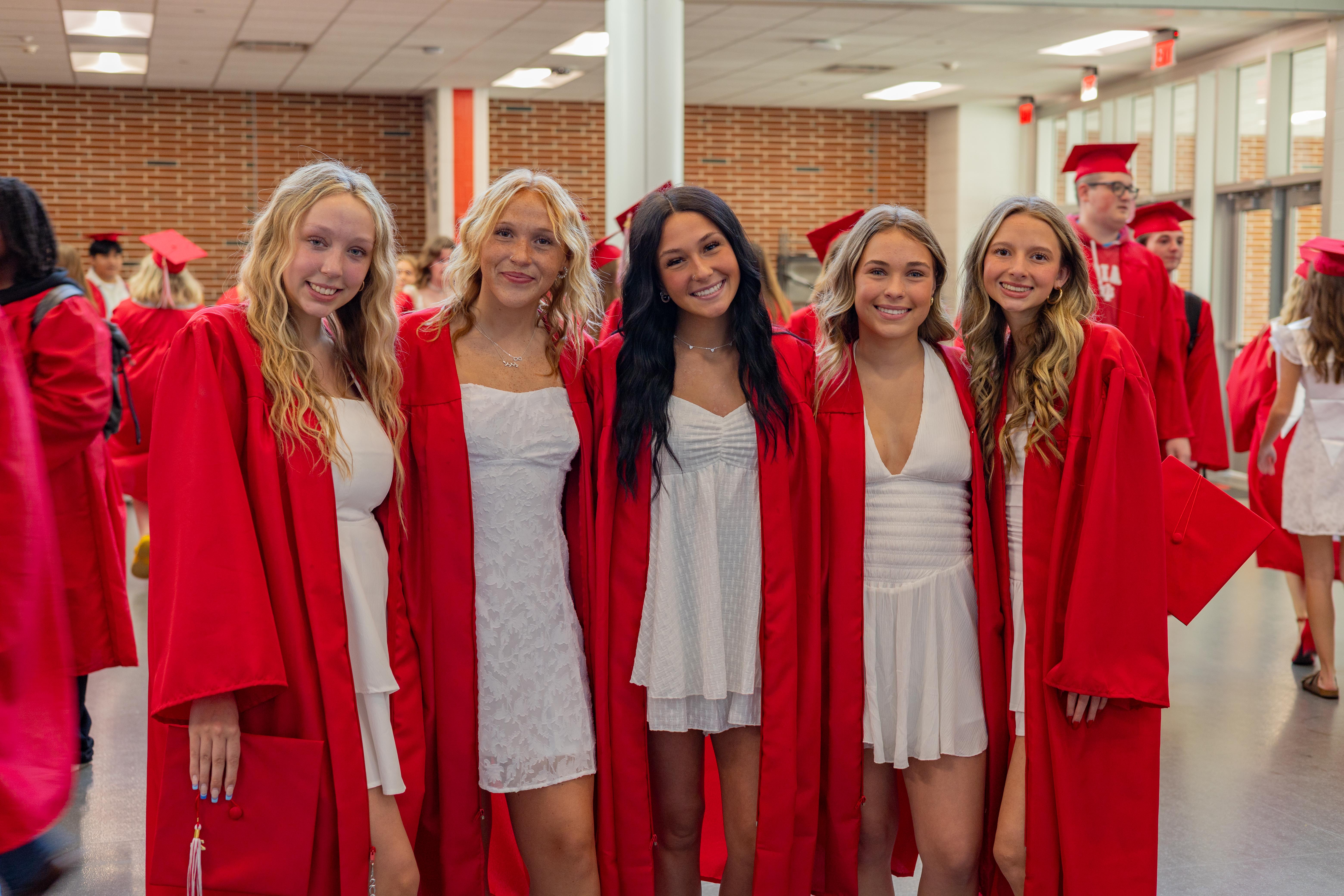Five smiling young women in white dresses and red graduation gowns stand in the foreground, while other graduates in similar attire and a hallway with windows are visible in the background.