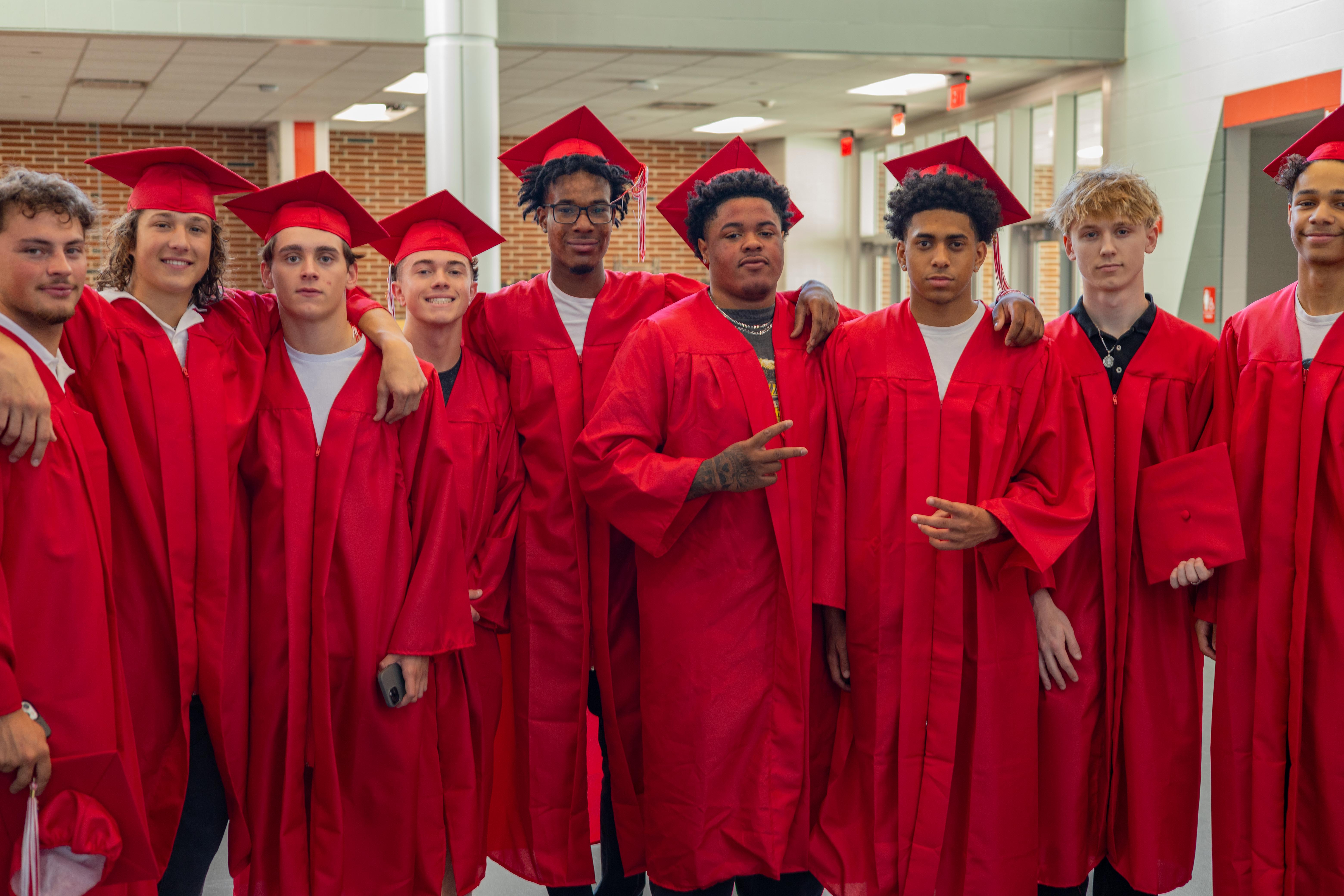 A group of young men in red graduation gowns and caps stand together in a hallway with lockers in the background.
