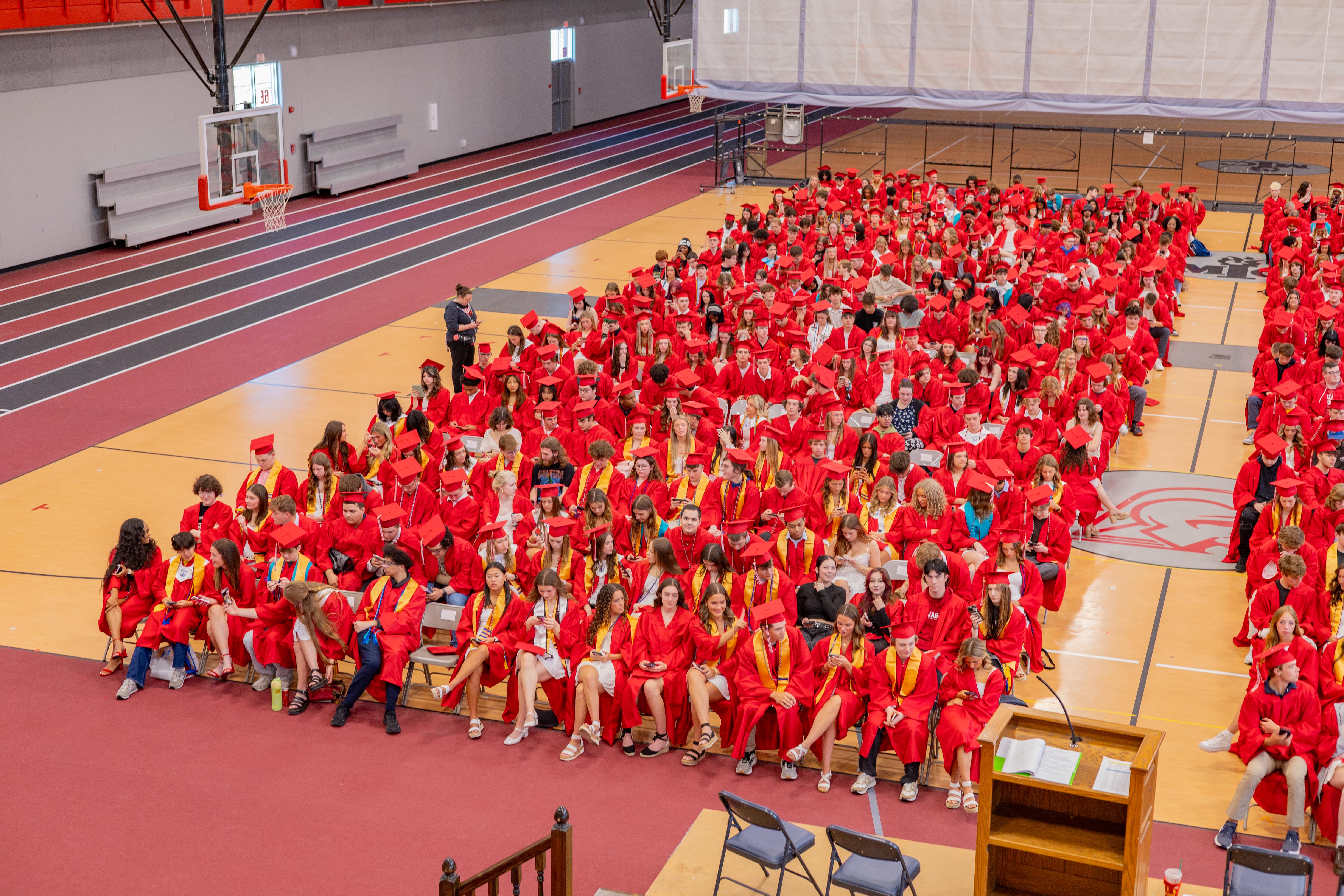 A large group of graduates in red gowns fill a gymnasium with bleachers and a track visible in the background.