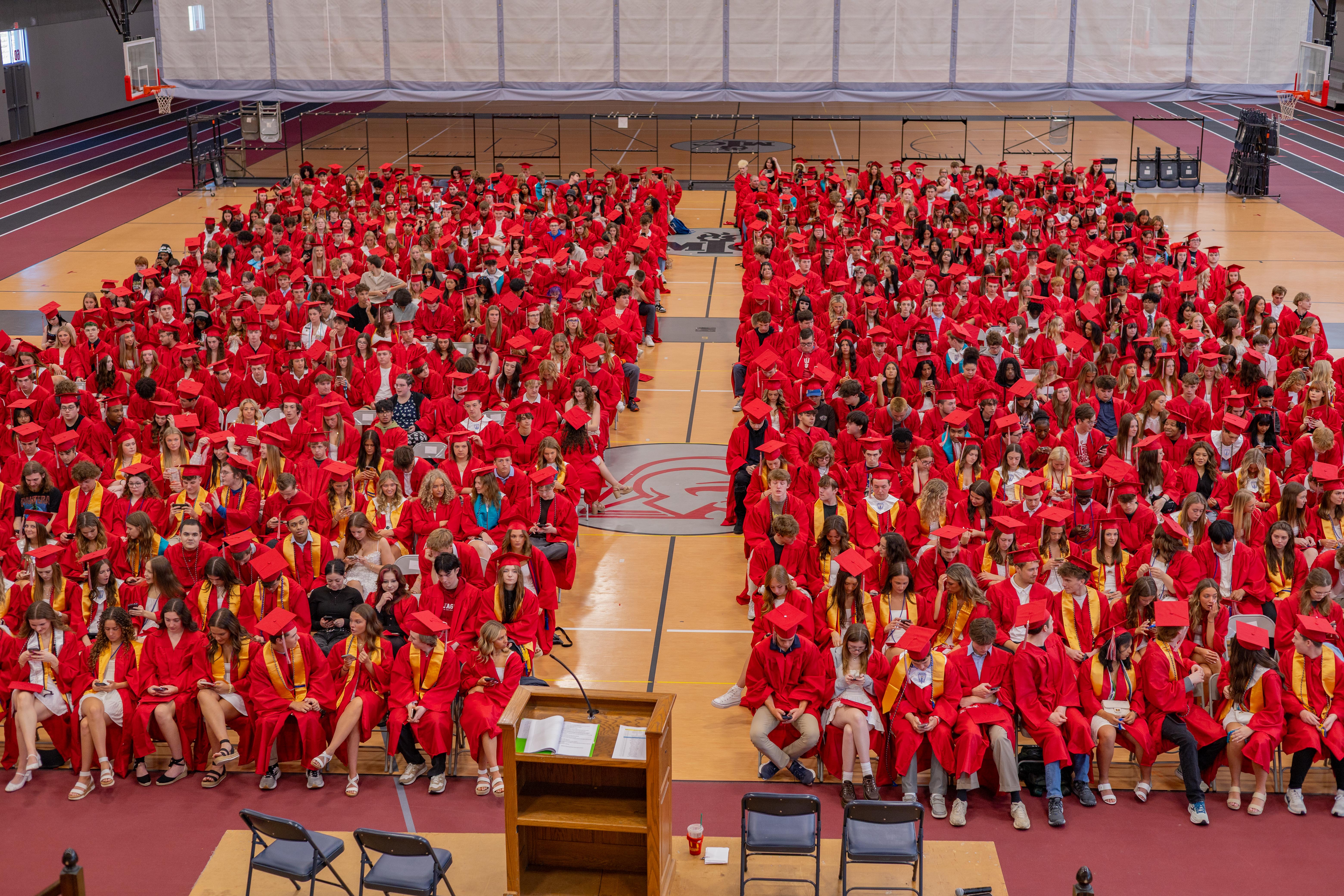 A large group of graduates in red gowns fill a gymnasium, with bleachers visible in the background.
