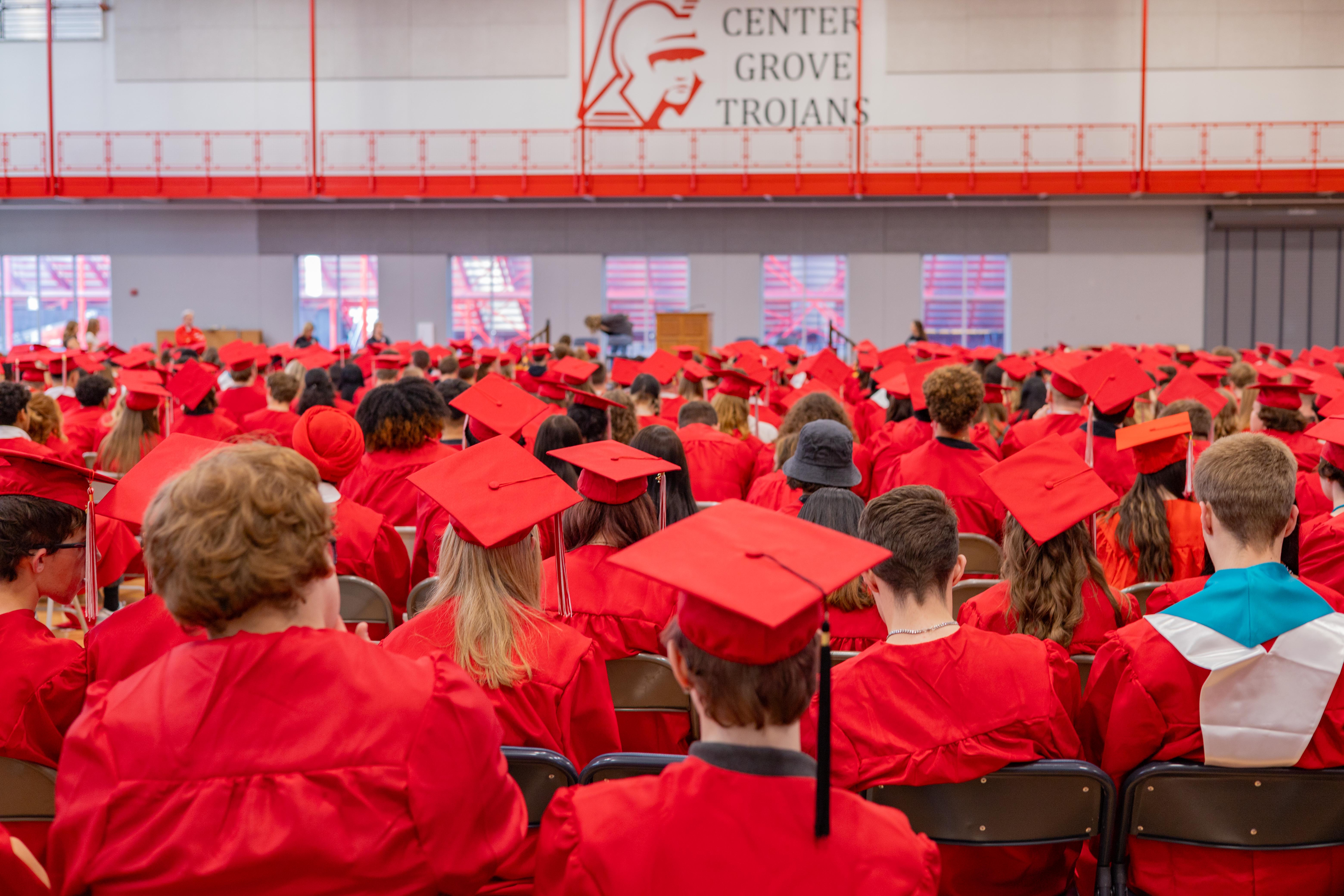 Graduates in red gowns and caps fill the foreground, facing a stage with a banner reading "CENTER GROVE TROJANS" in the background.