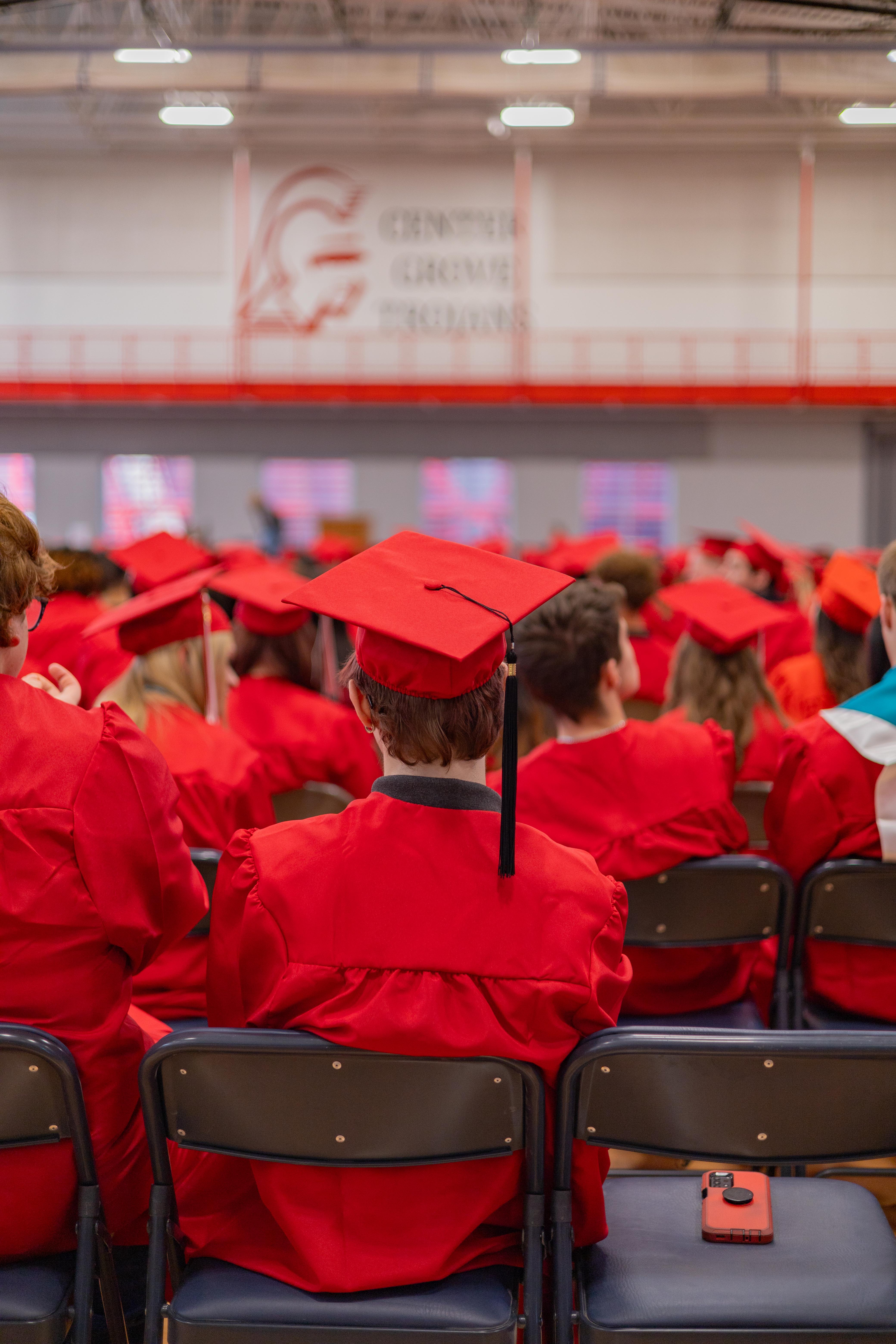 Graduates in red gowns and caps sit in chairs in the foreground, facing a large banner with a red logo in the background.