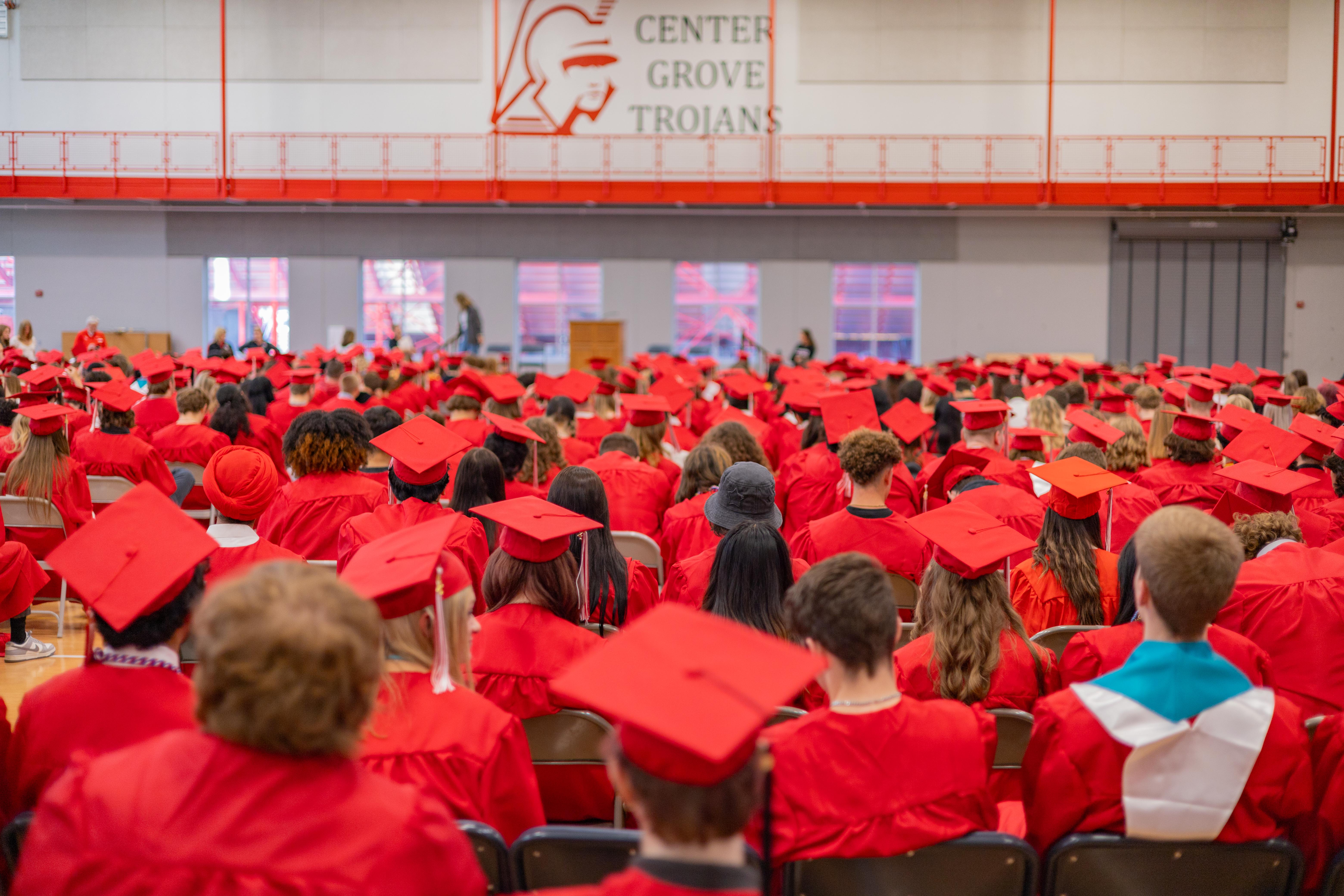 A large group of graduates in red gowns and caps are seated in an auditorium with a banner that reads "CENTER GROVE TROJANS" in the background.