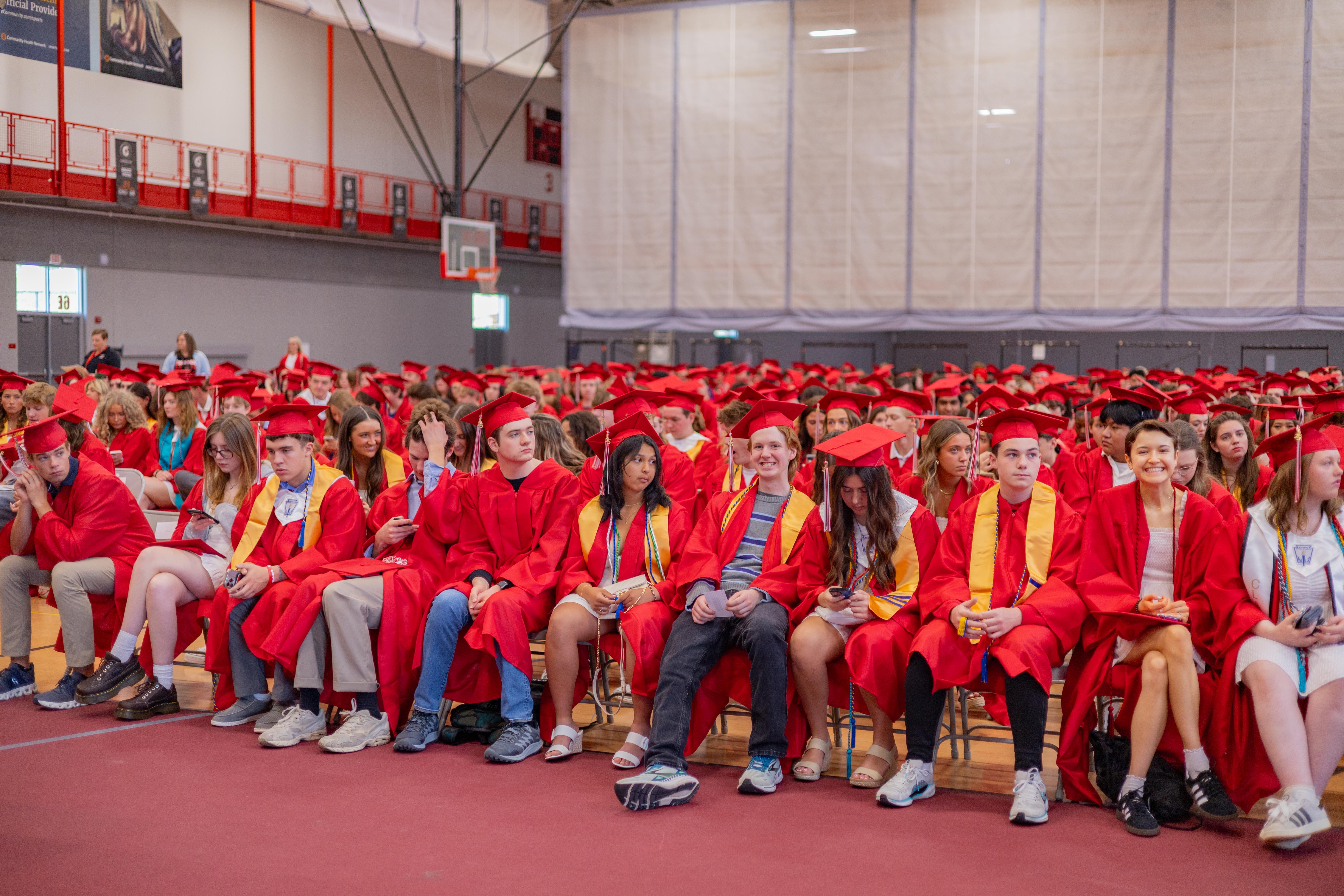 A group of graduates in red gowns and caps sit in a gymnasium, with bleachers and a basketball hoop visible in the background.