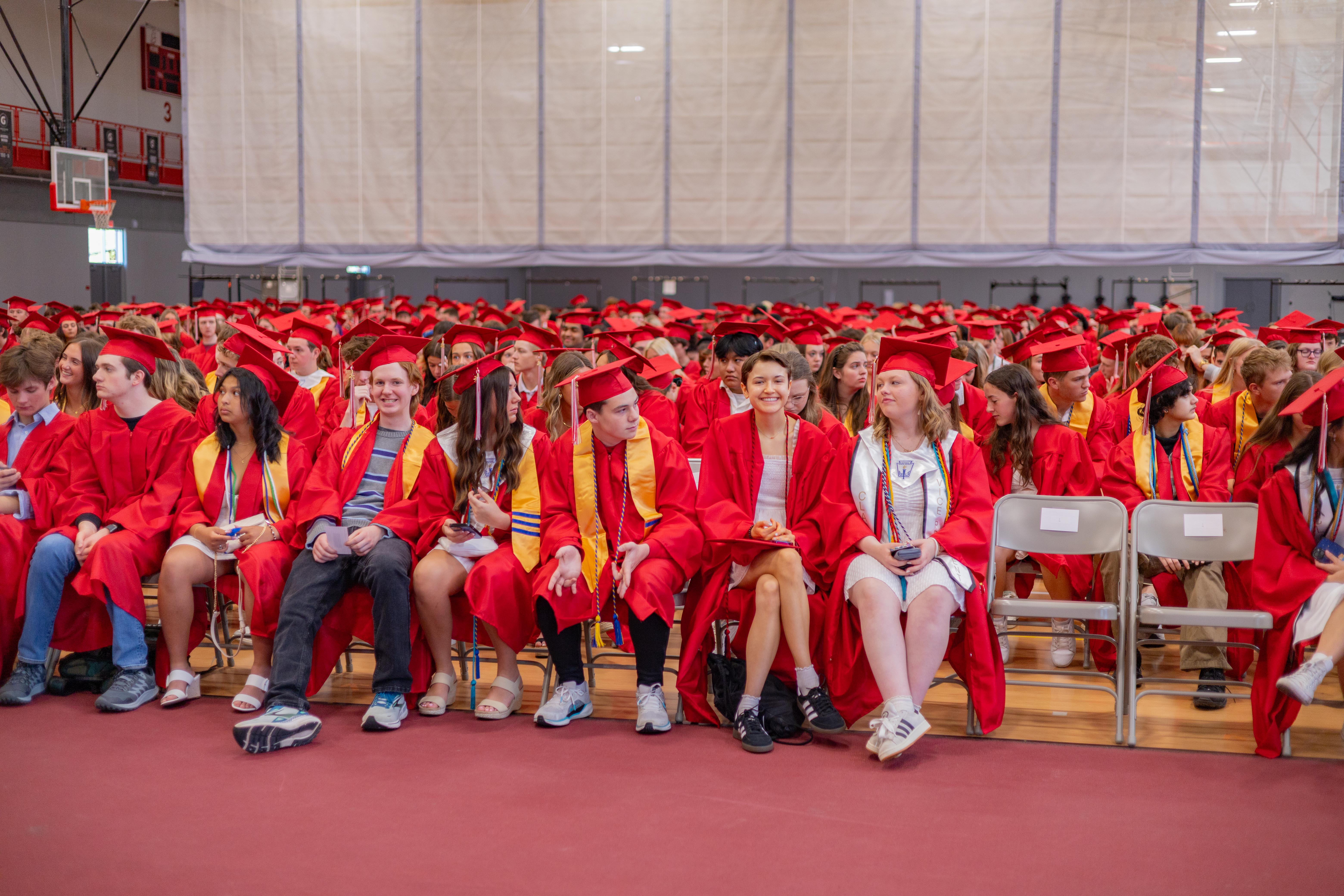 A large group of graduates in red gowns and caps sit in a gymnasium with a red floor in the foreground and bleachers visible in the background.
