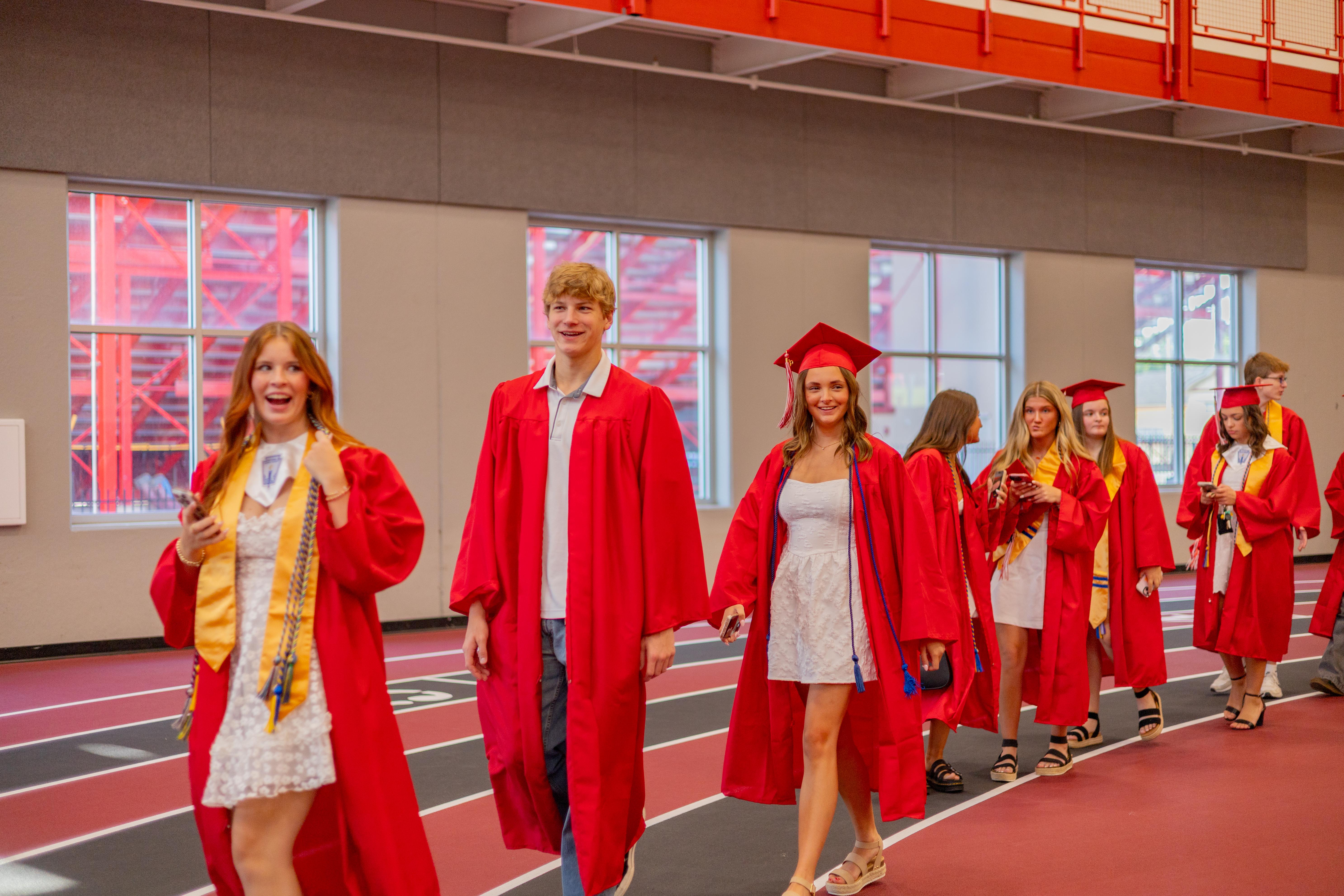 A group of graduates in red gowns and caps walk down a track, with bleachers visible in the background.