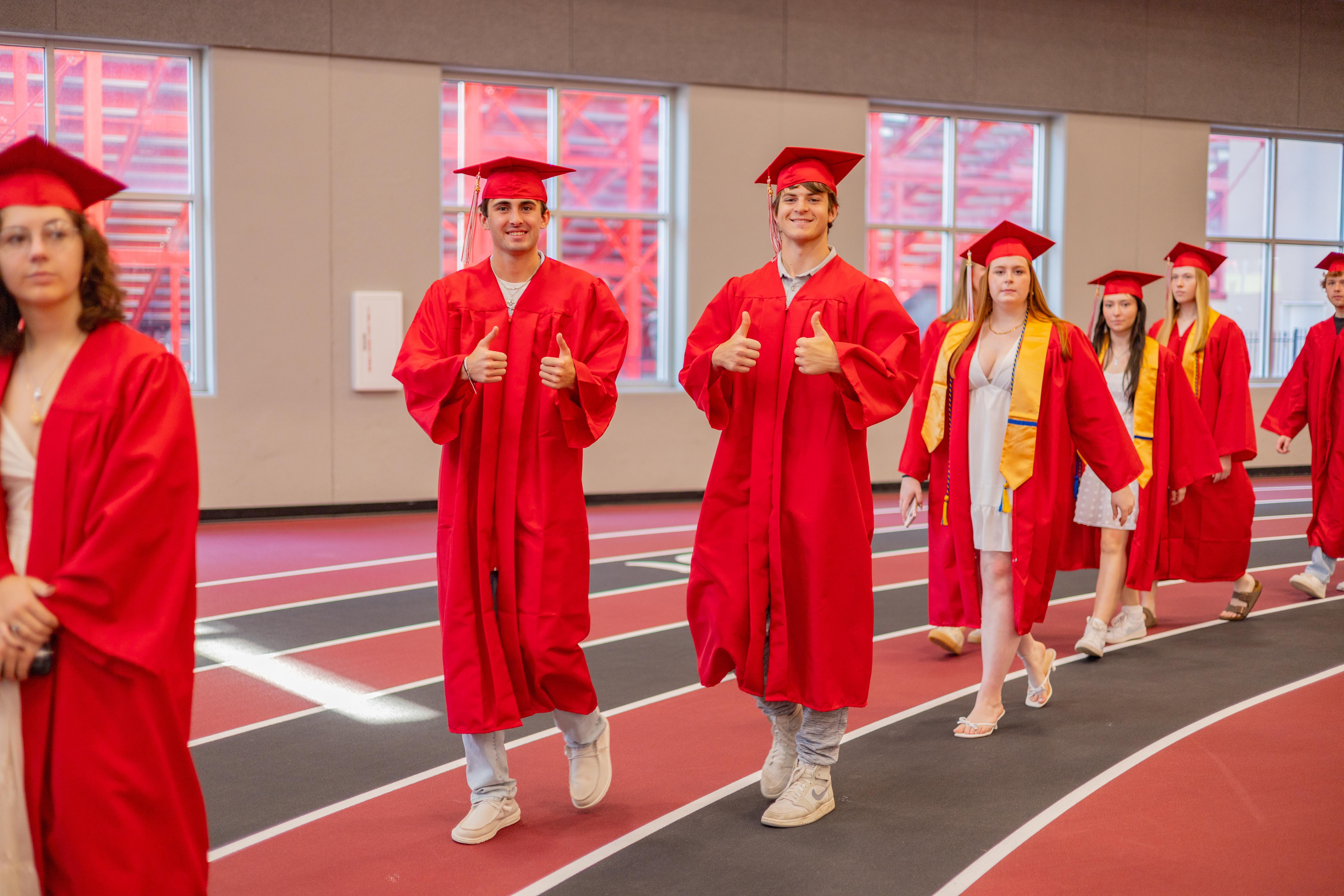 A group of graduates in red gowns and caps walk down a track, with large windows and red scaffolding visible in the background.