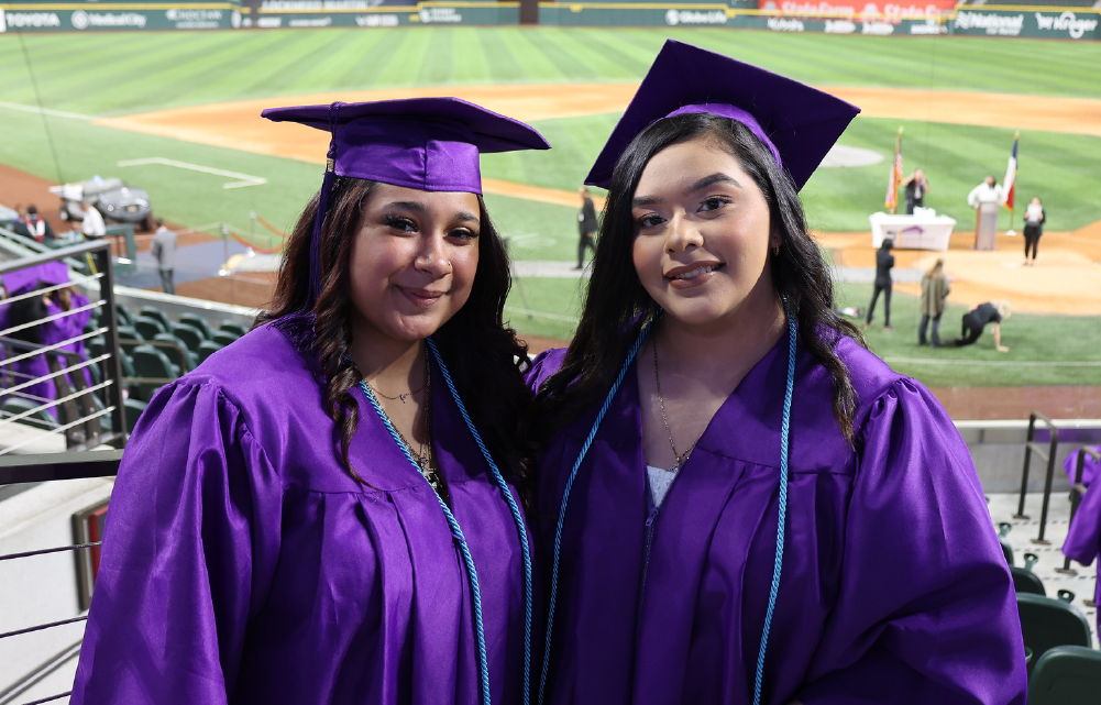 Two young women in purple graduation gowns and caps stand together in front of a baseball stadium.