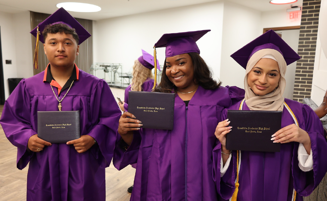 Three individuals in purple graduation gowns and caps stand together, holding their diplomas, in what appears to be a hallway or lobby area.