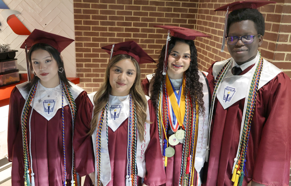 Four individuals wearing graduation gowns and caps stand together in front of a brick wall, celebrating their academic achievement.